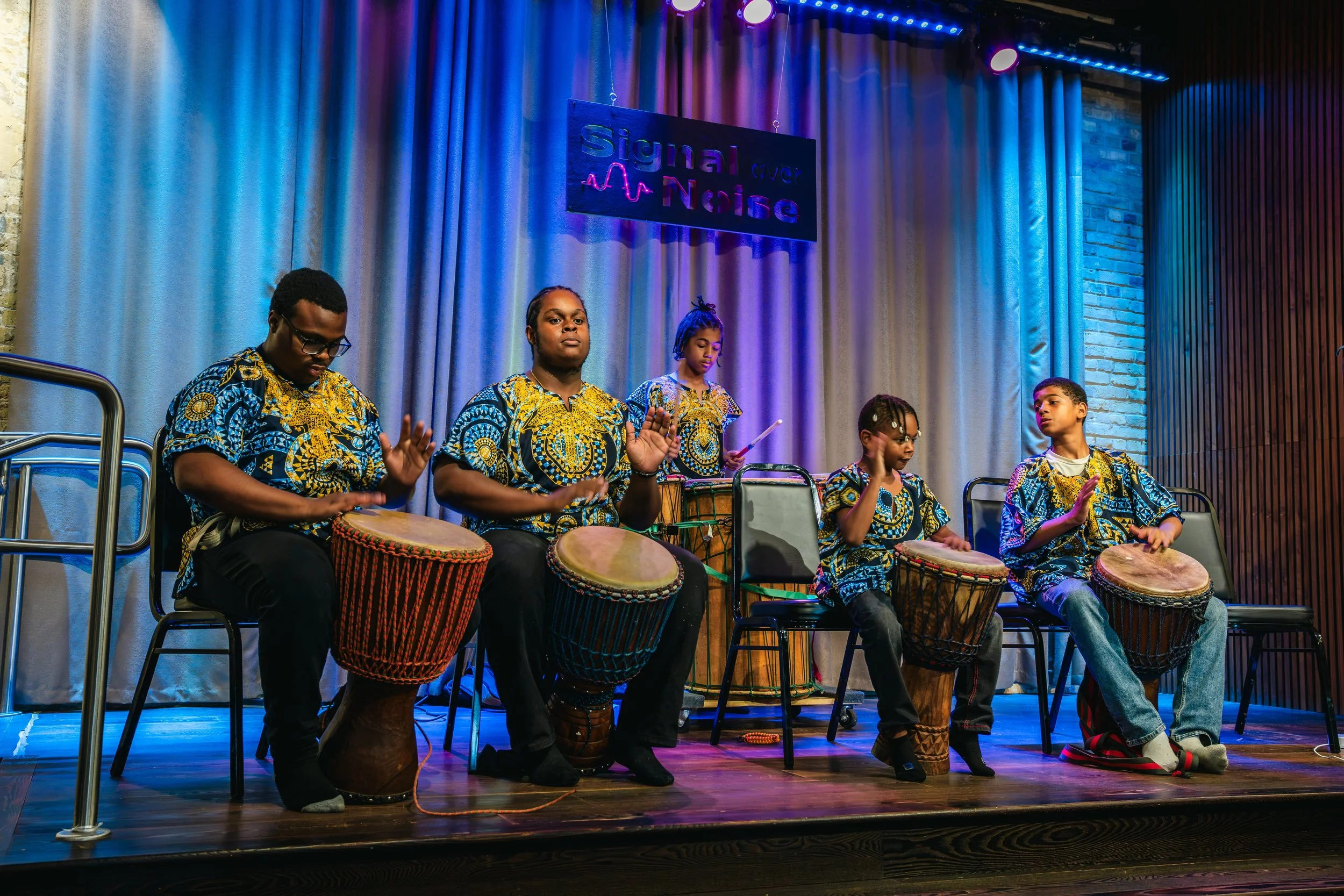 A group of children playing various drums on stage during a performance. They are dressed in colorful African-style clothing. The stage has a curtain backdrop with a sign that reads 'Signal Over Noise' and colorful stage lighting.
