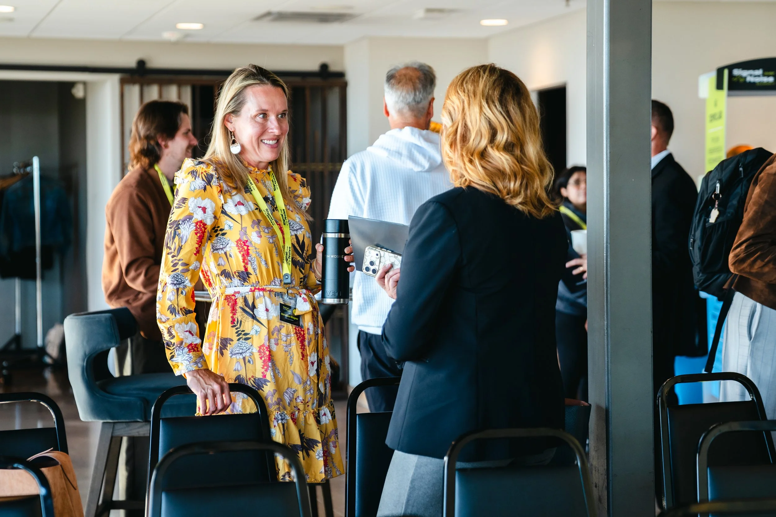 People gathered at an indoor event, engaging in conversation. A woman in a yellow floral dress is smiling while talking to another woman with blonde hair. Multiple chairs are arranged around and several attendees are in the background.