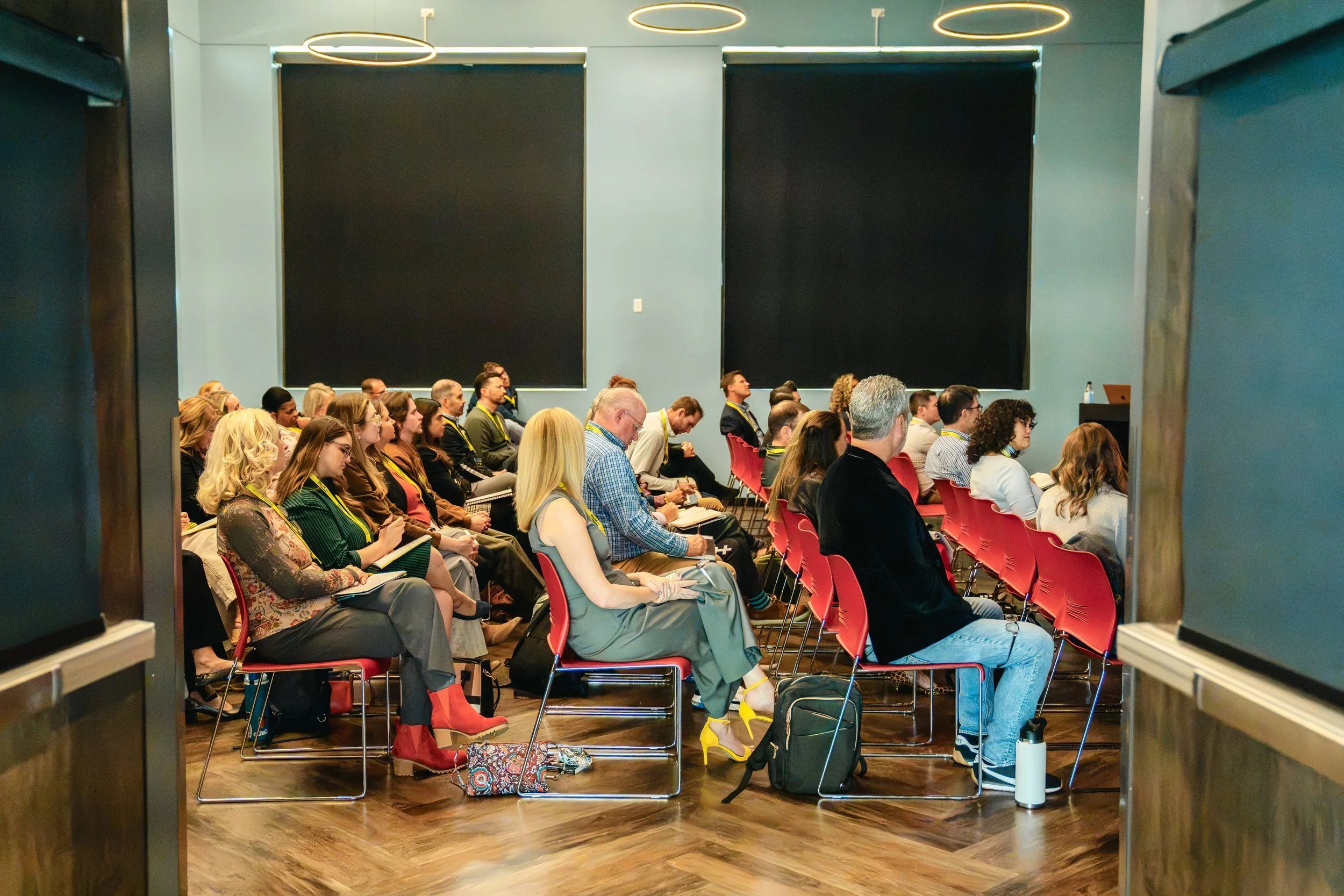 People seated in rows attending a conference or seminar, viewed through a doorway with black panels and circular ceiling lights.