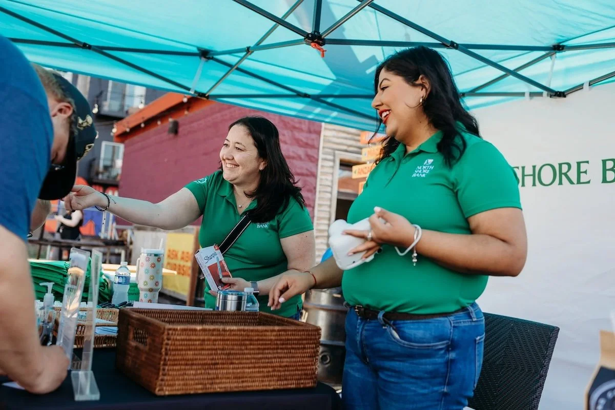 Two women in green shirts smiling and engaging with a person at a booth under a teal canopy at an outdoor event.