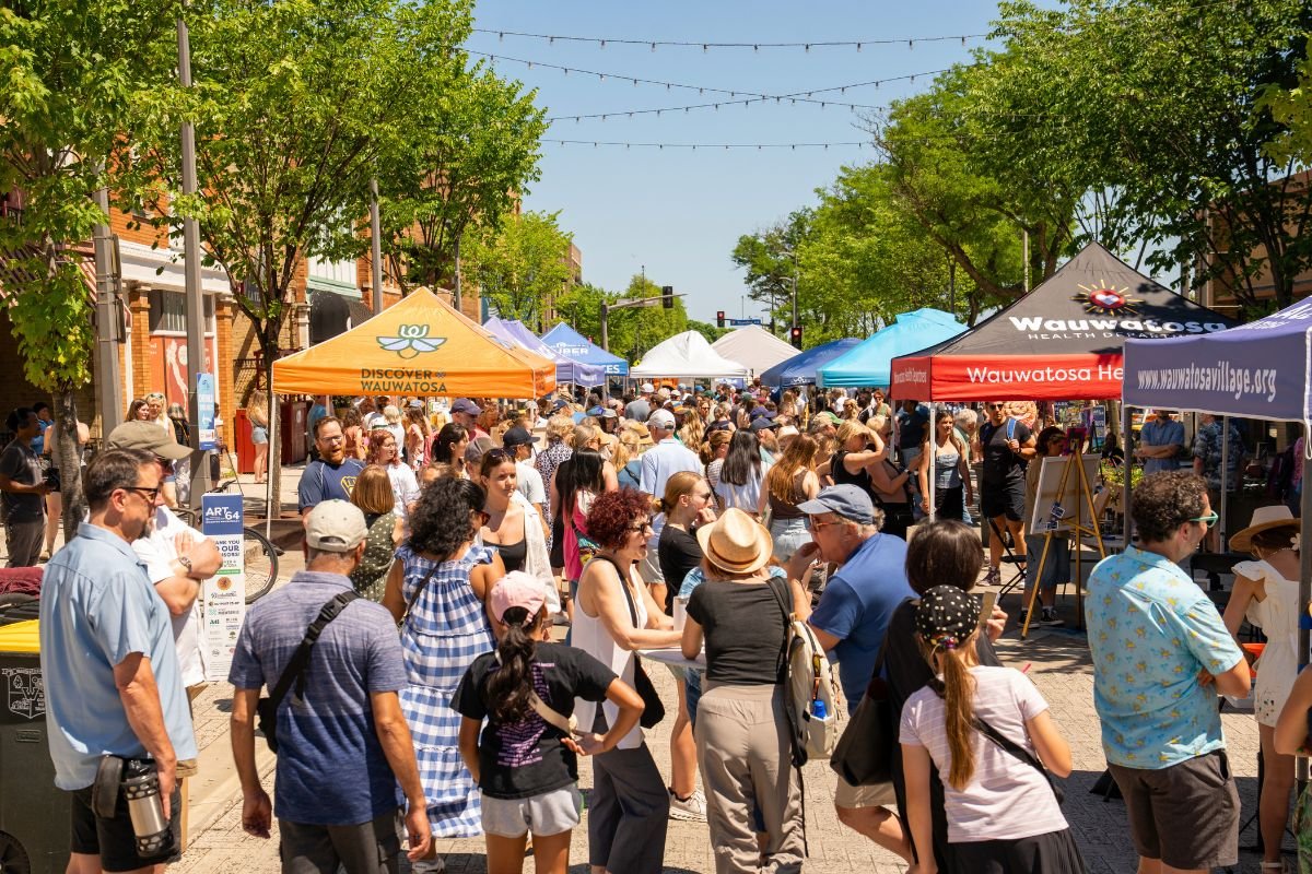 Crowd of people at an outdoor street event with colorful vendor tents, trees, and string lights overhead.