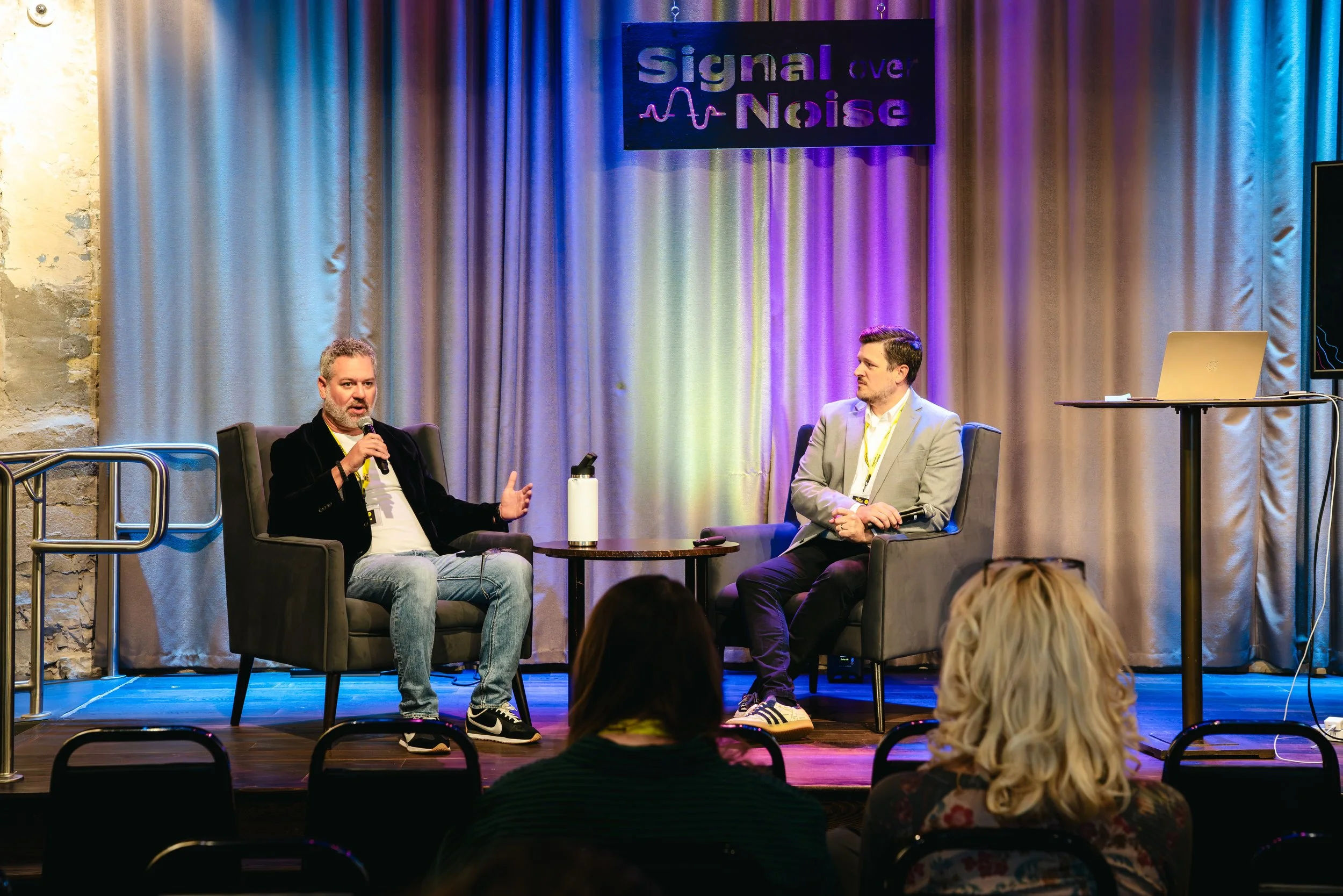 Two men are sitting on stage in conversation during a panel discussion at an event called 'Signal over Noise.' One man is speaking into a microphone, while the other listens. The stage background features curtains and a sign with the event name. Audience members are watching in front of the stage.