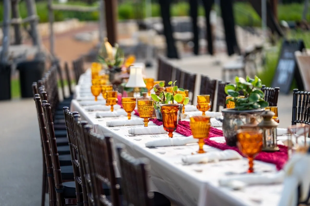 A long outdoor dining table set with orange glassware, white napkins, and potted plants, with chairs on each side and a blurred background of greenery.