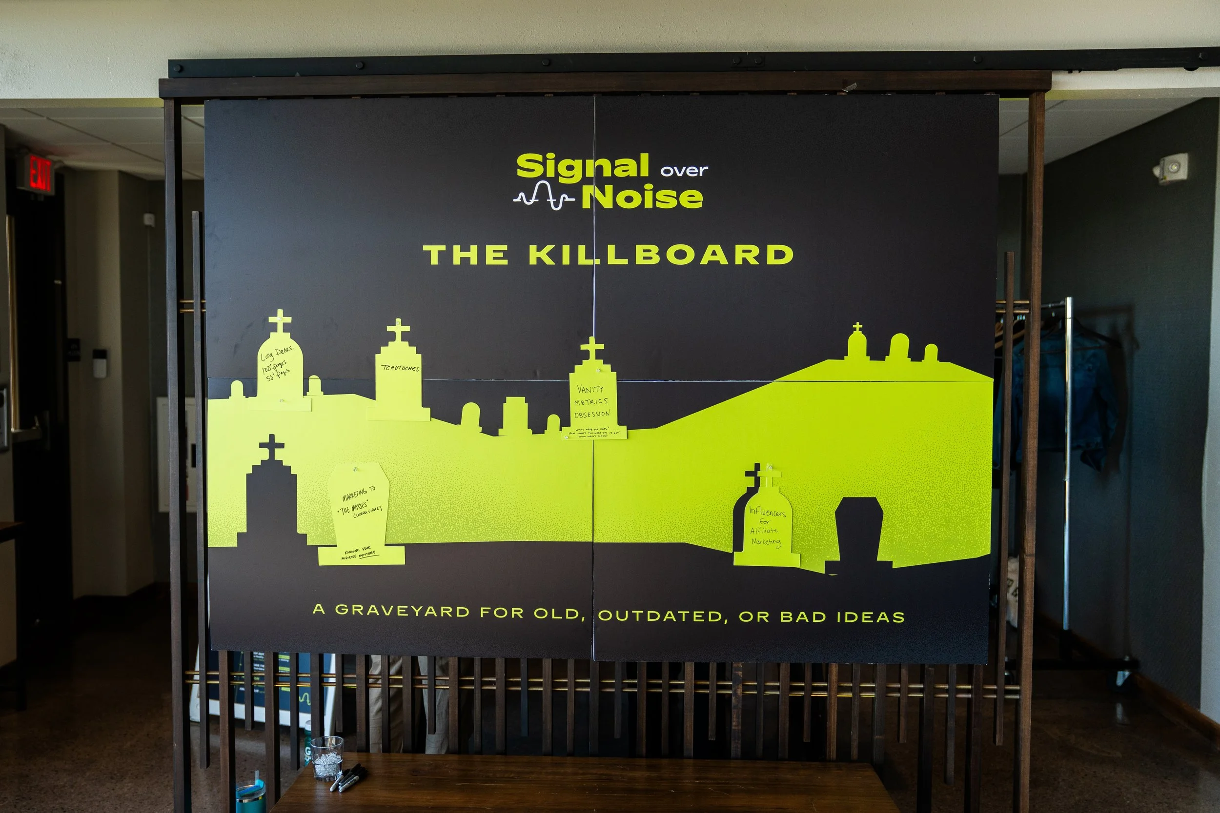 Poster with the title 'Signal over Noise' and subtitle 'The Killboard.' The poster features a city skyline silhouette with church steeples and buildings, and has several yellow sticky notes attached with handwritten notes. The bottom of the poster reads 'A Graveyard for Old, Outdated, or Bad Ideas.' The setting appears to be an indoor space, possibly a conference or workshop.
