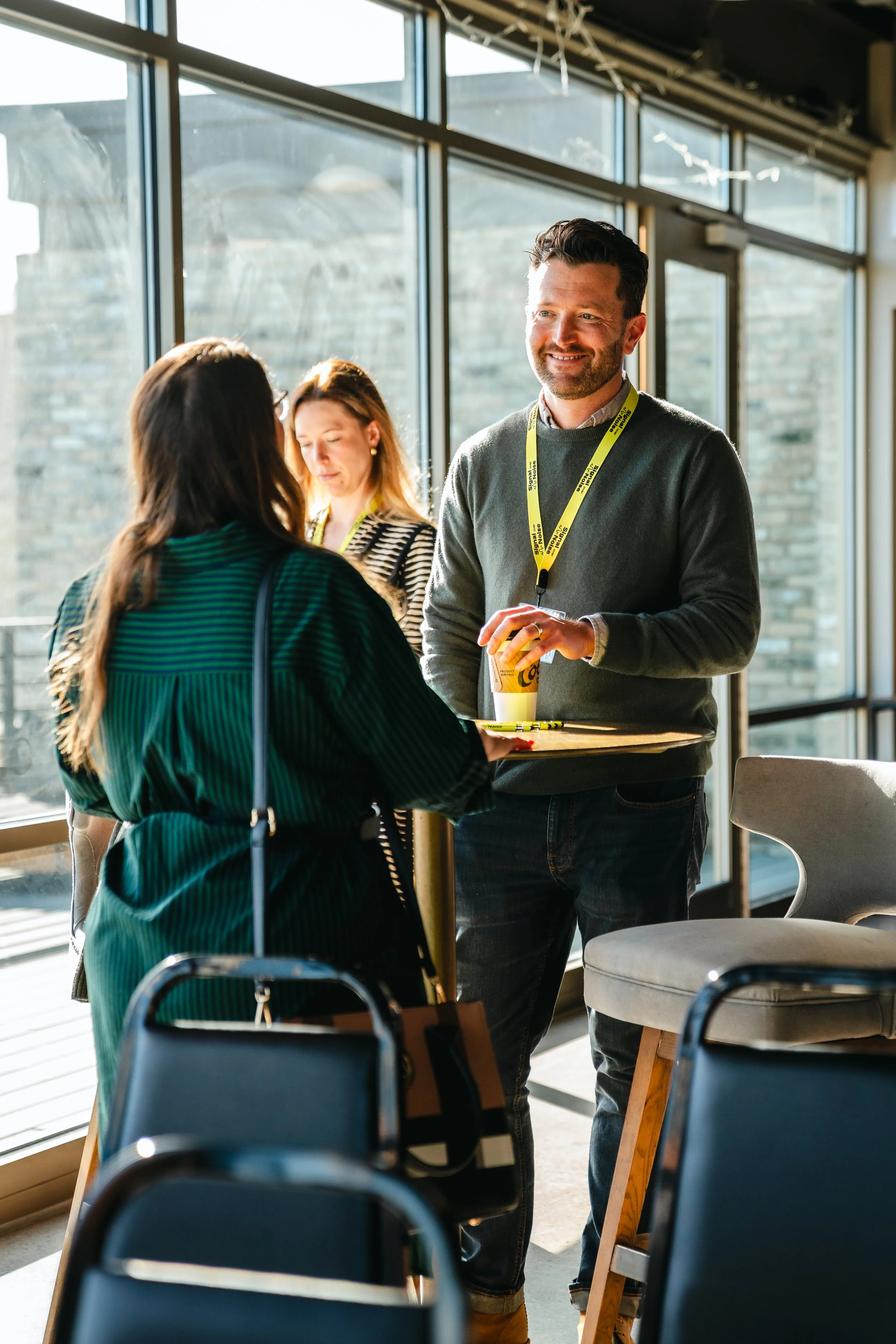 People having conversations in a brightly lit indoor setting with large windows, casual attire, and yellow lanyards.