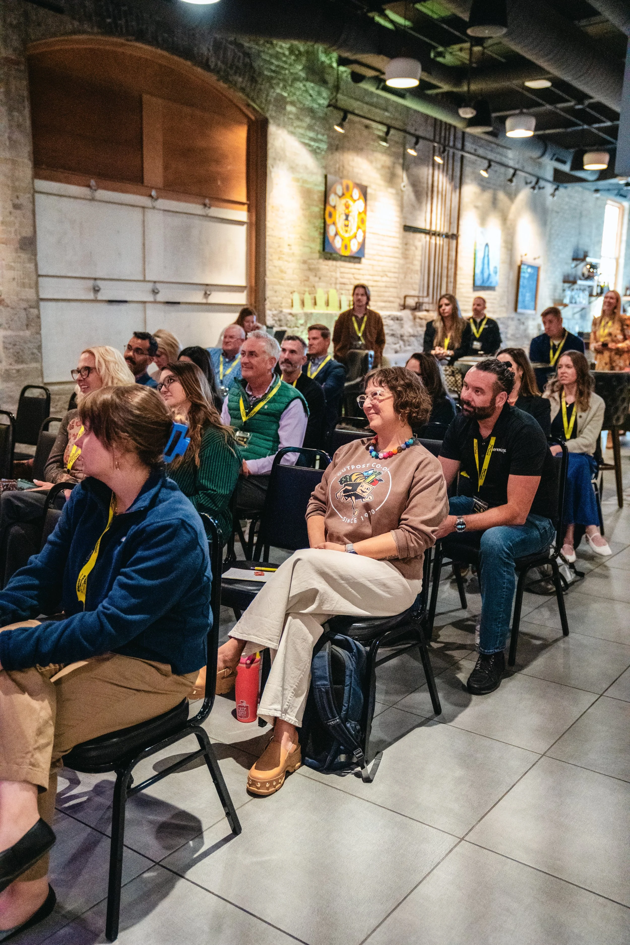 People attending a conference or seminar, sitting in a dimly lit room with brick walls, and art pieces hanging on the wall, wearing yellow lanyards.