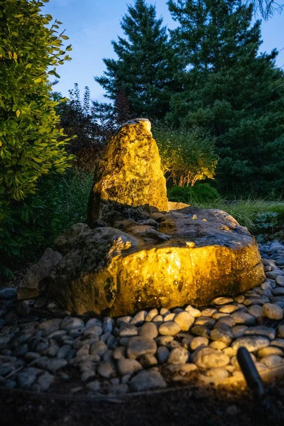 Decorative outdoor garden fountain with a large illuminated stone centerpiece surrounded by small pebbles, greenery, and tall trees at dusk.