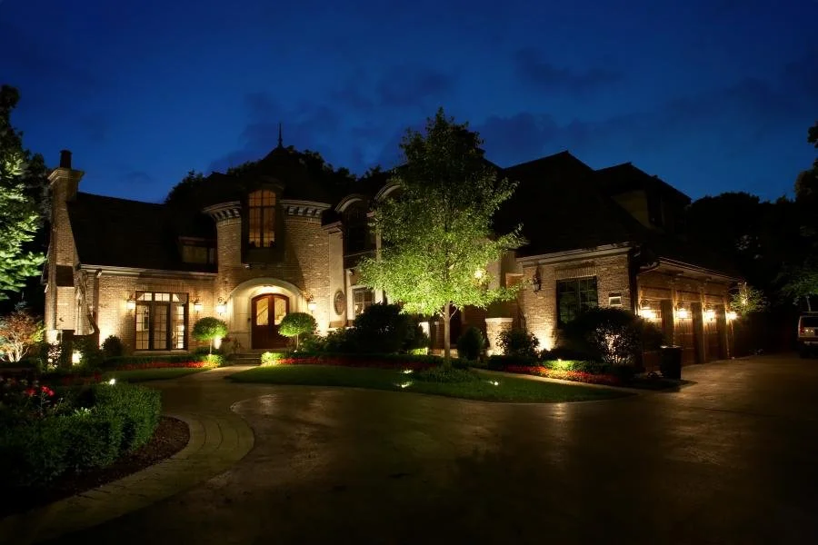 Large, elegant brick house illuminated at night with well-lit landscaping, driveway, and tall trees.
