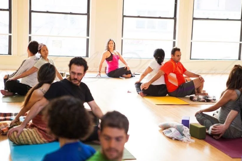 Group of people participating in a yoga class in a spacious room with large windows, sitting on yoga mats in a cross-legged position.