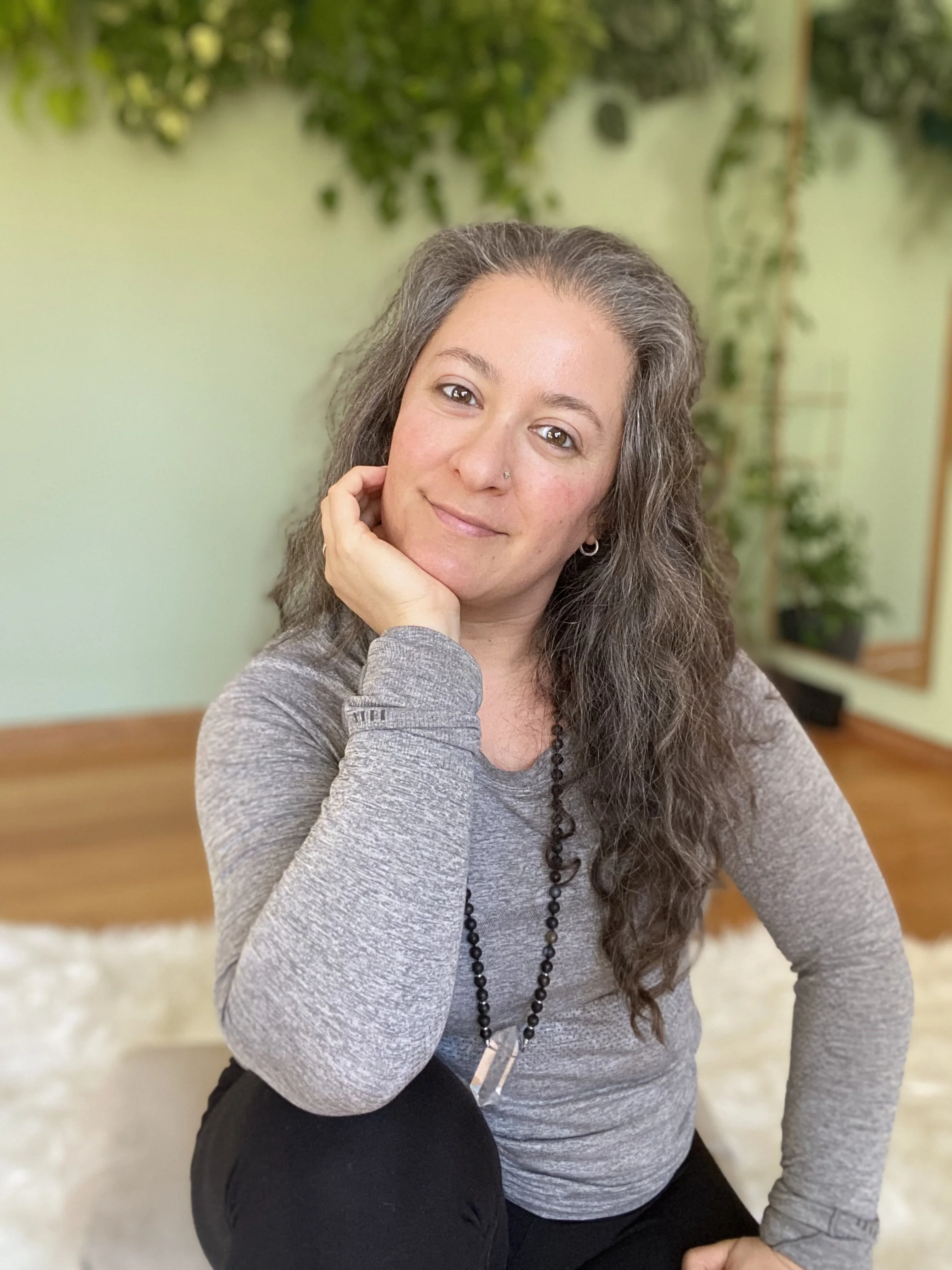 A woman with curly dark hair and a nose piercing sits on the floor, smiling softly at the camera, with a green background of blurred plants.