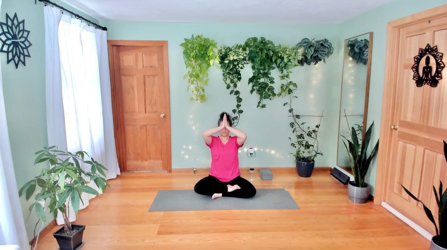 A woman practicing yoga in a bright, airy room with wooden floors and green walls adorned with potted plants and string lights. She is sitting cross-legged on a yoga mat, holding her hands in prayer position overhead.