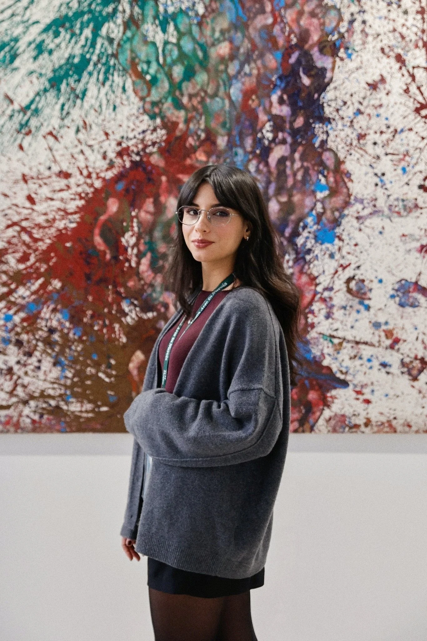 Donna con capelli scuri e occhiali a cerchio, indossa un maglione grigio e una gonna nera, si trova davanti a un dipinto astratto colorato.