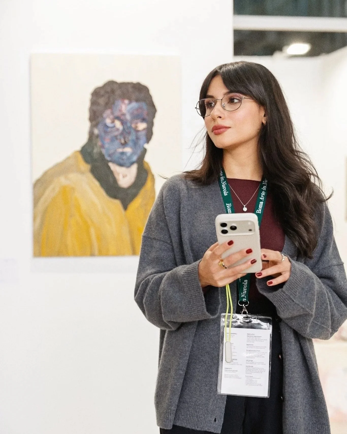 Donna con occhiali, capelli scuri e vestito scuro osserva un'opera d'arte in un museo, indossa un badge e tiene in mano uno smartphone.