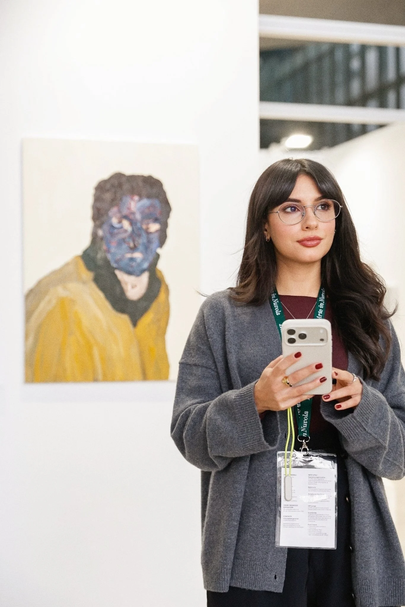 Social Media Manager per l'arte con capelli castani, occhiali e giacca grigia, che tiene in mano uno smartphone, si trova in una mostra d'arte con un quadro di un uomo con maglietta gialla alle spalle.
