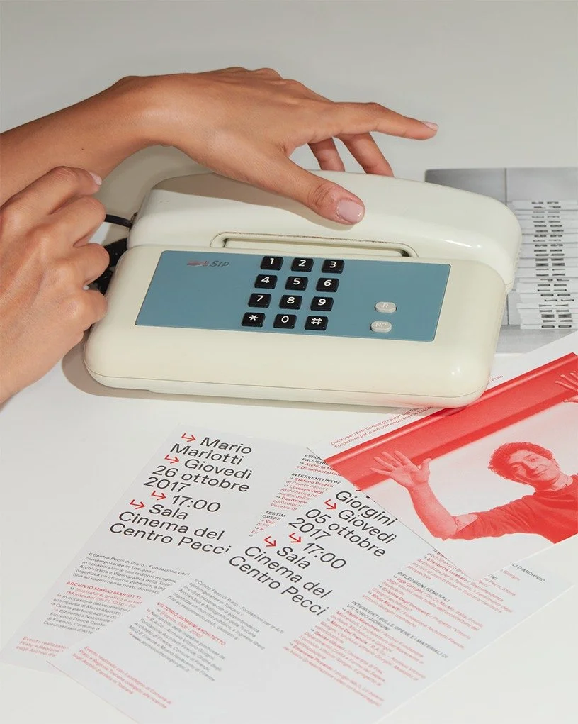 Telefono a rotella bianco sopra una scrivania, accanto a fogli di carta con testi e un biglietto di invito a un evento al cinema.