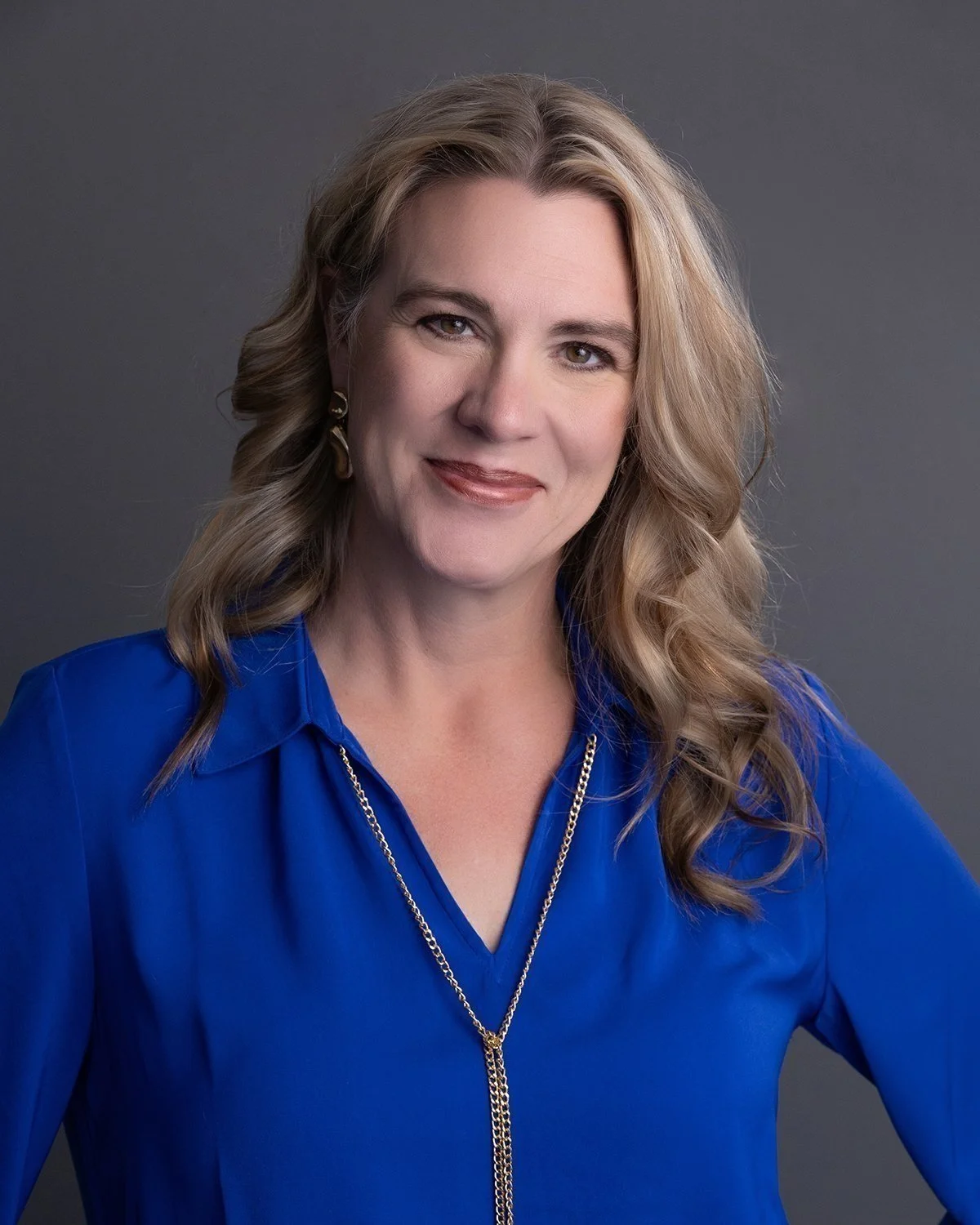 A woman with long, wavy blonde hair smiling at the camera, wearing a bright blue top, gold earrings, and a gold necklace against a plain gray background.