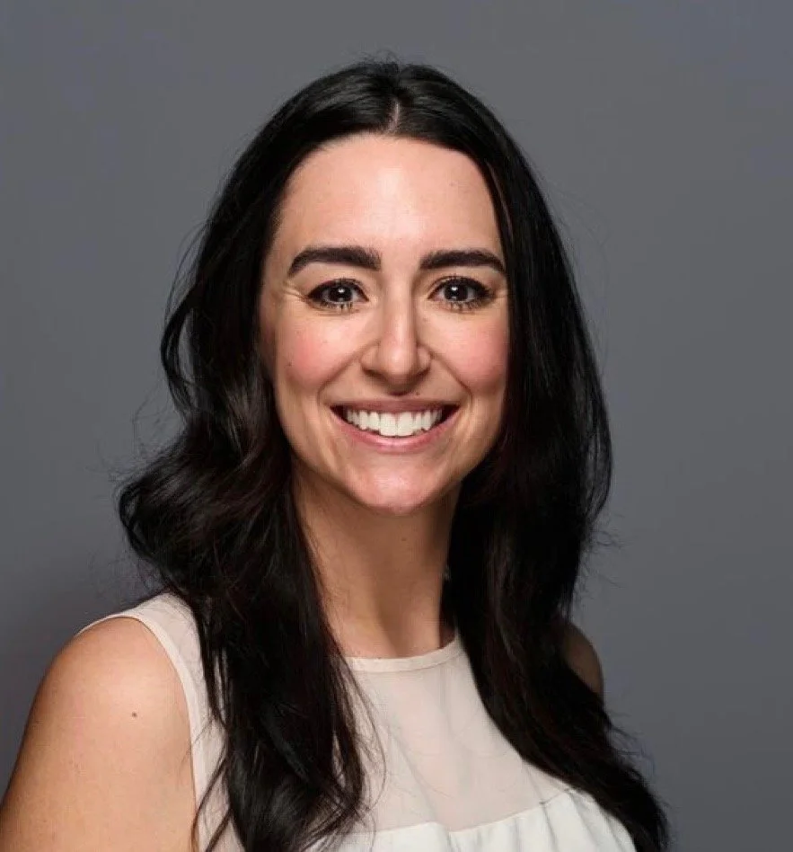 A woman with long dark hair and a big smile, wearing a sleeveless light-colored top, against a plain gray background.