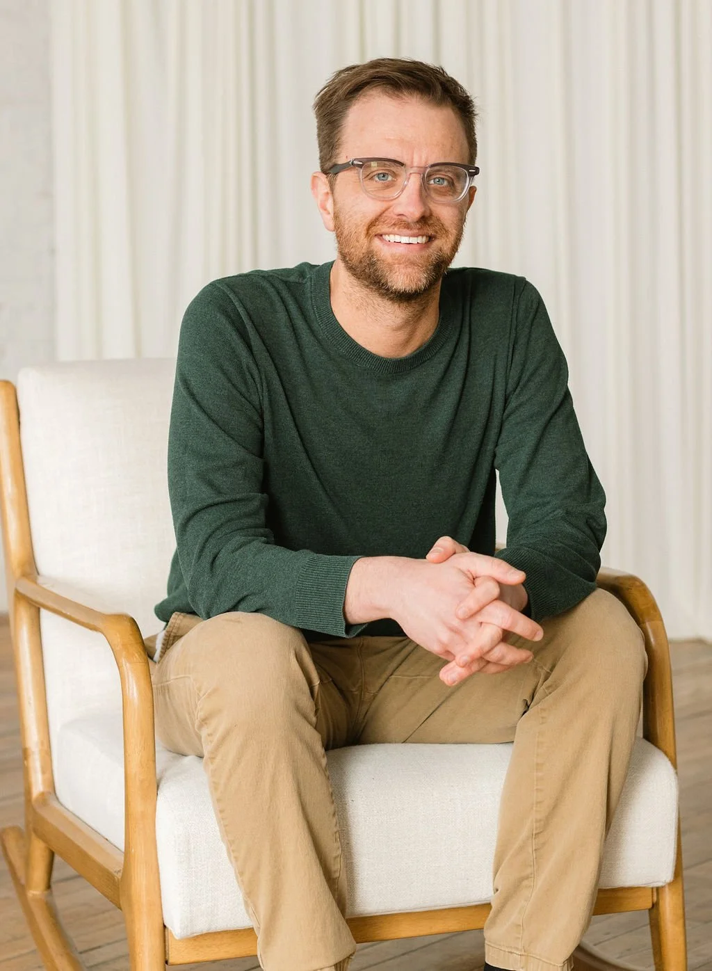 A man with glasses and a beard sitting on a white cushion armchair, smiling, wearing a dark green sweater and tan pants, with a light-colored curtain background.