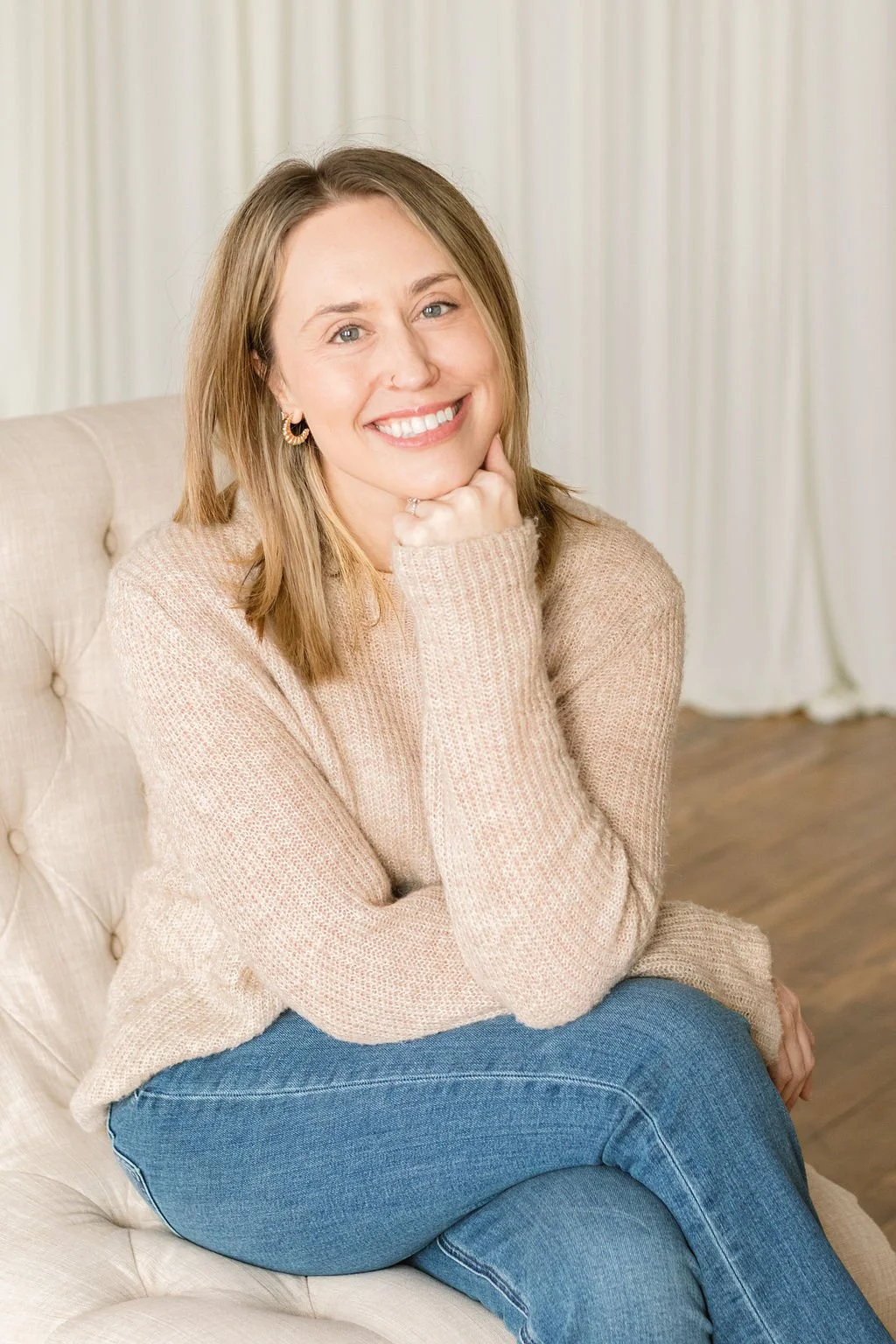A woman with shoulder-length light brown hair and blue eyes smiling, sitting on a beige sofa with her hand resting on her chin, wearing a beige knit sweater and blue jeans, in a bright room with white curtains.