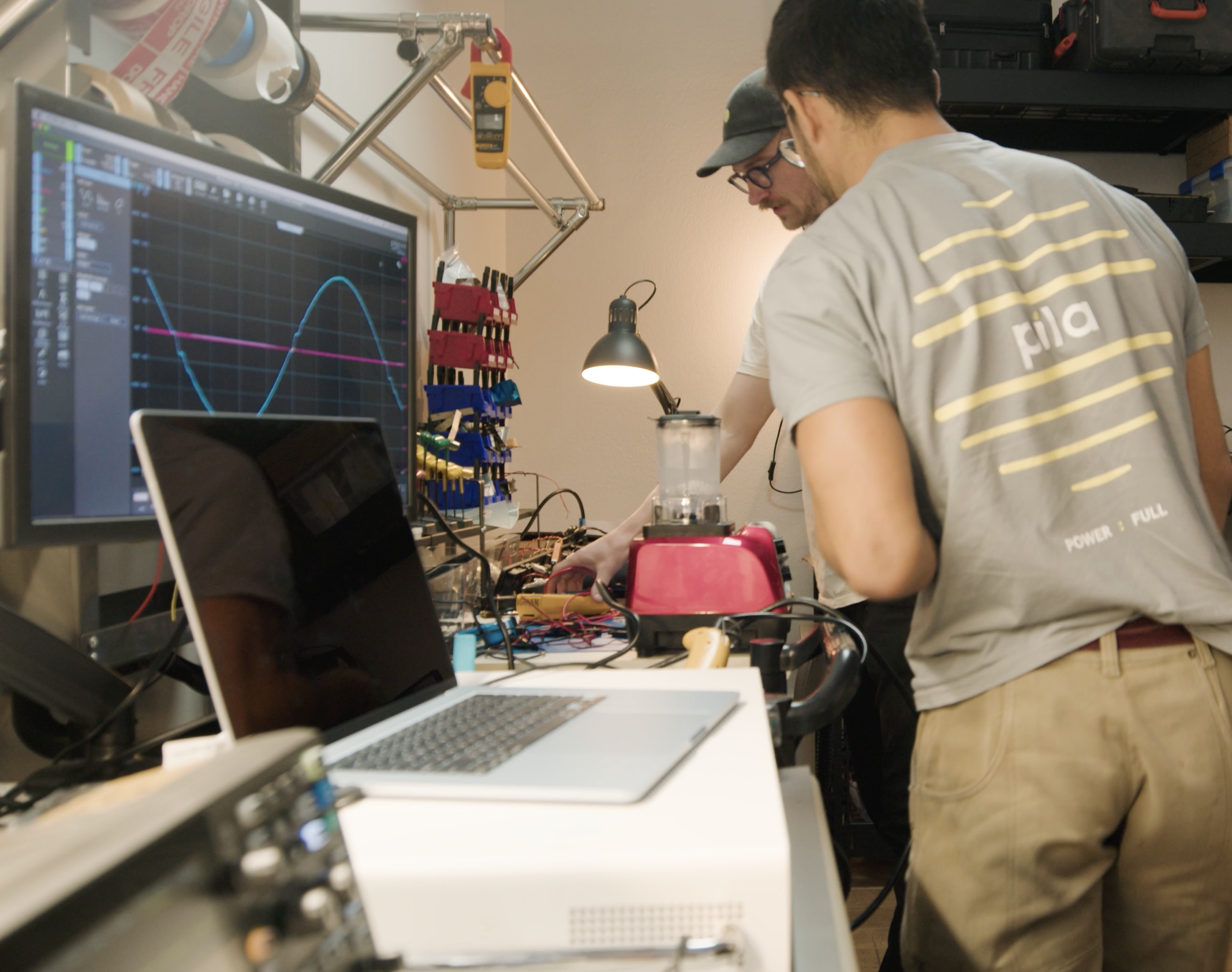 Two people working on electronics experiments at a lab bench, with a computer monitor displaying waveforms, various tools, and electronic components around them.