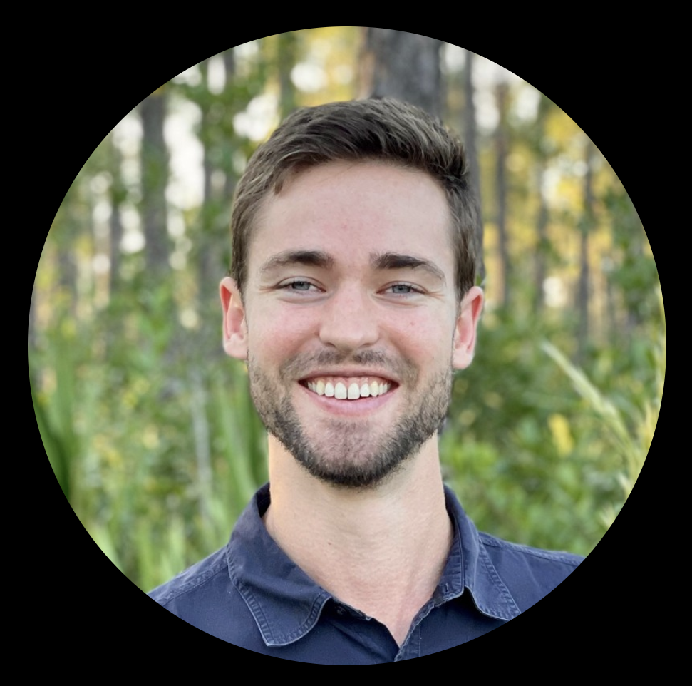 A young man with short brown hair and a beard, smiling with teeth showing, in an outdoor setting with lush green plants and trees in the background.