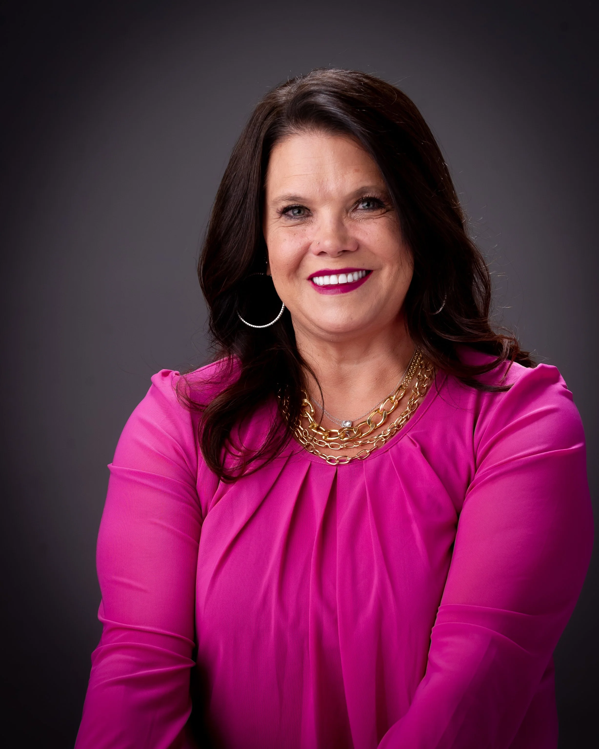 A woman with dark brown hair, wearing a bright pink long-sleeve blouse, layered gold necklaces, hoop earrings, and dark lipstick, smiling in front of a dark gray background.