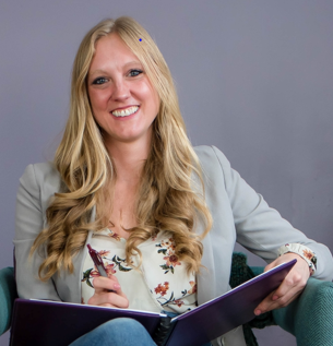 A woman with long blonde hair, smiling, sitting on a teal chair, holding a purple notebook and pen, in front of a gray background.