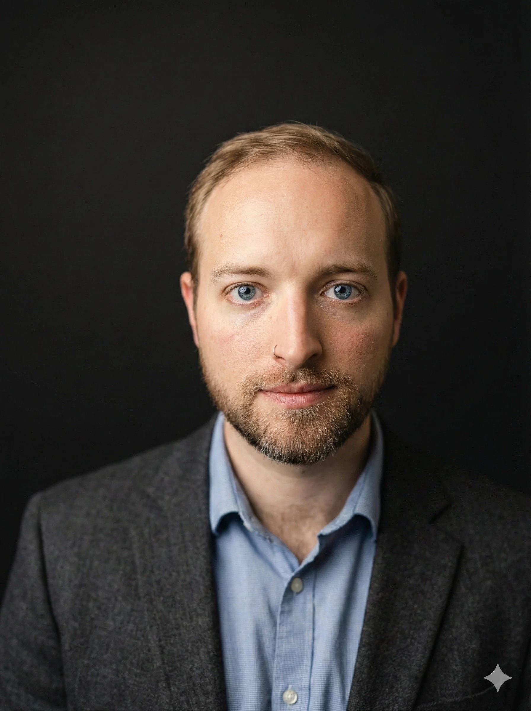 A young man with short brown hair, wearing a blue short-sleeve shirt and tattoos on his arms, stands outdoors by a river with crossed arms, under a clear blue sky, and trees in the background.