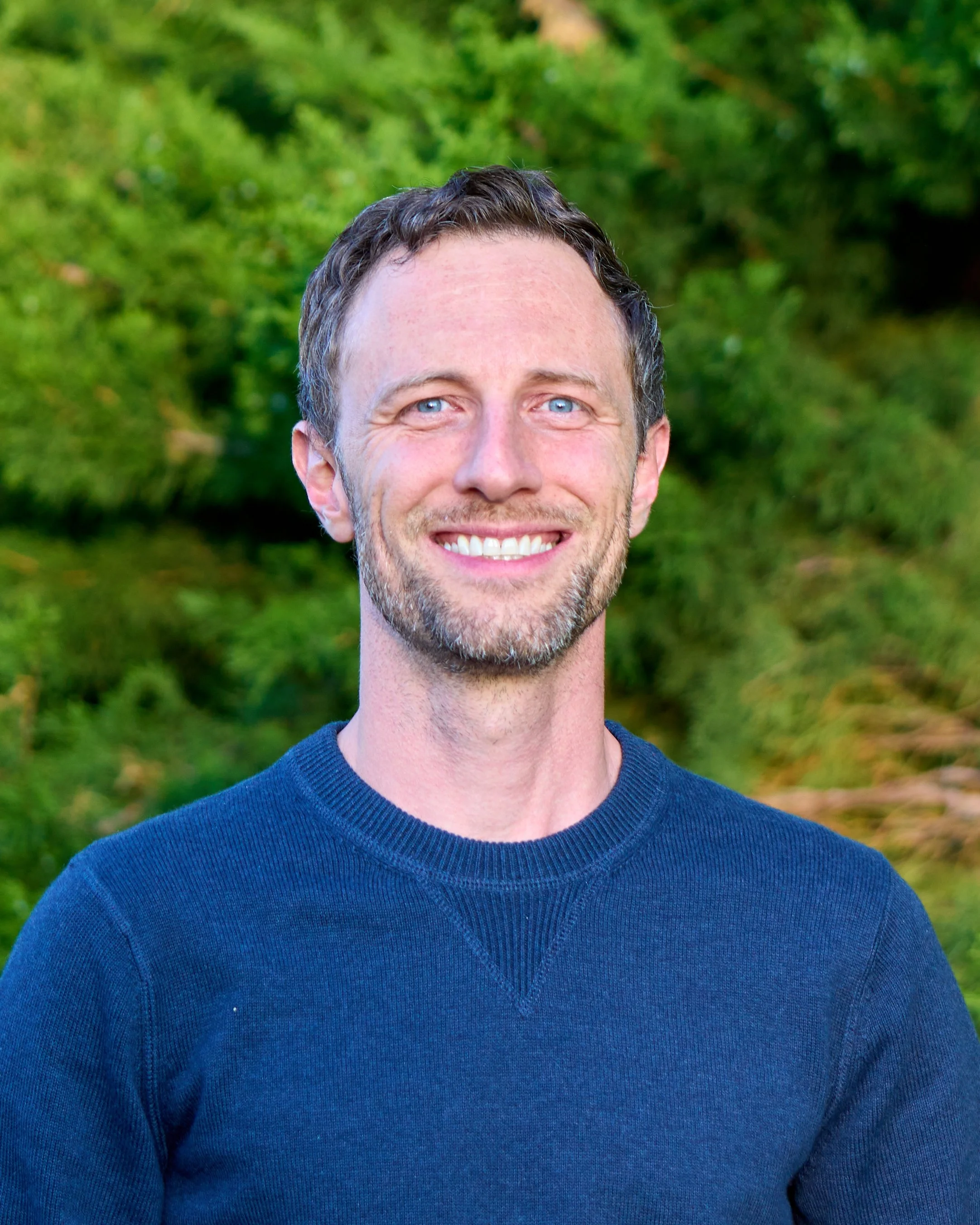 A man with short dark hair, blue eyes, and a beard smiling outdoors with a green, blurred background.