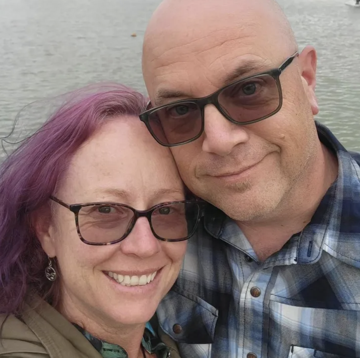 A smiling couple taking a selfie outdoors near a body of water, with the woman having purple hair and wearing glasses, and the man being bald, wearing glasses, and a plaid shirt.
