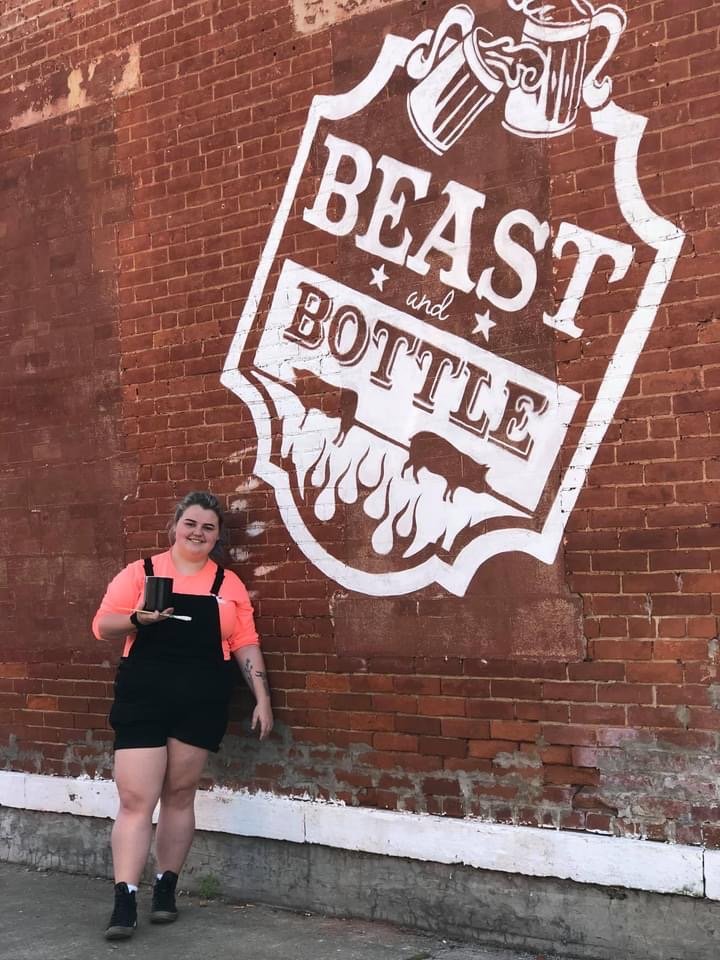 A woman standing in front of a brick wall with a large white mural that reads 'Beast and Bottle' with illustrations of beer mugs and flames.