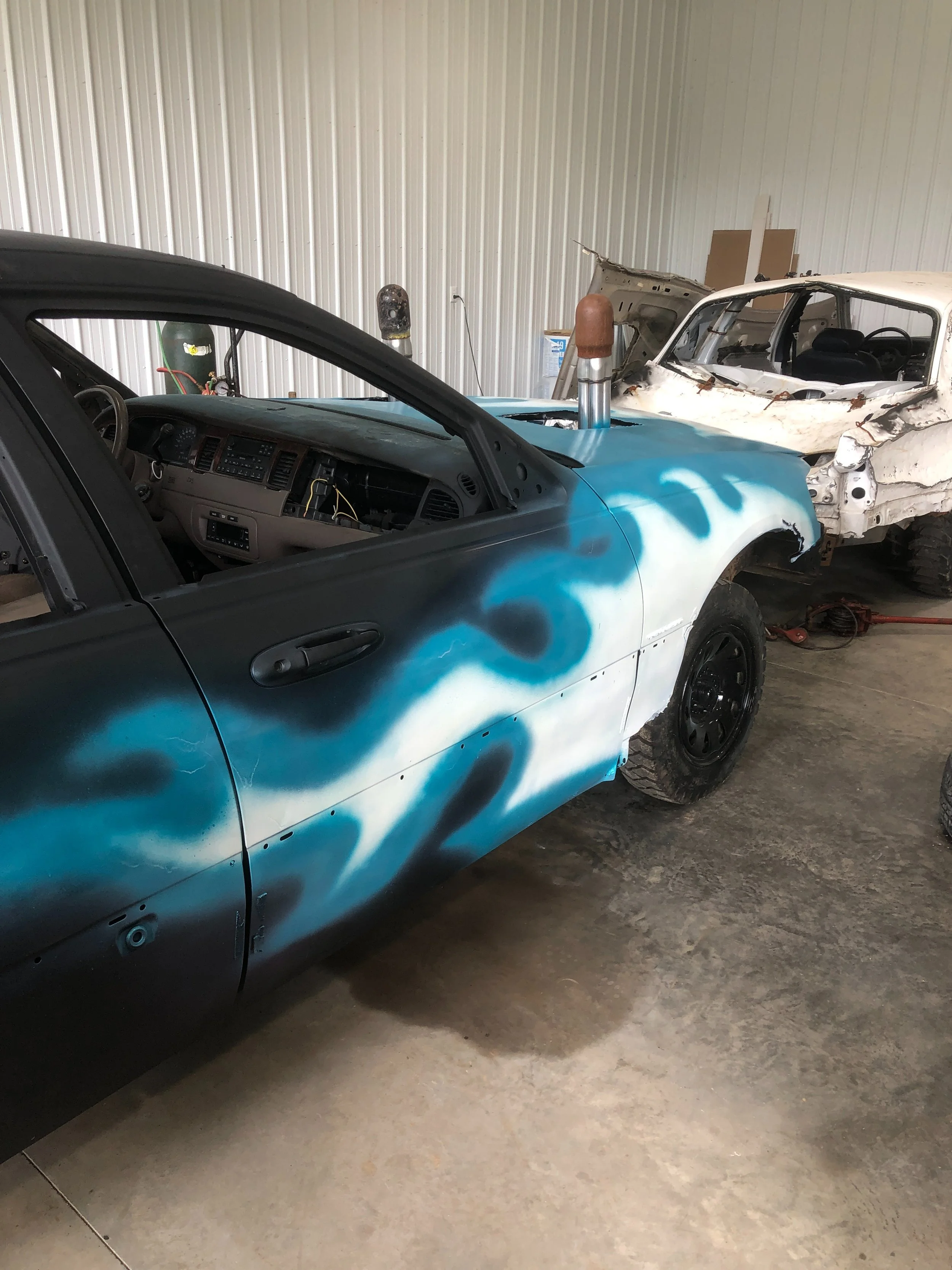 A garage with a black and blue spray-painted car in the foreground and a white, damaged car in the background. The black and blue car has a pop-up headlight, a modified or custom paint job with spray patterns, and a large pipe with a brown cap extend