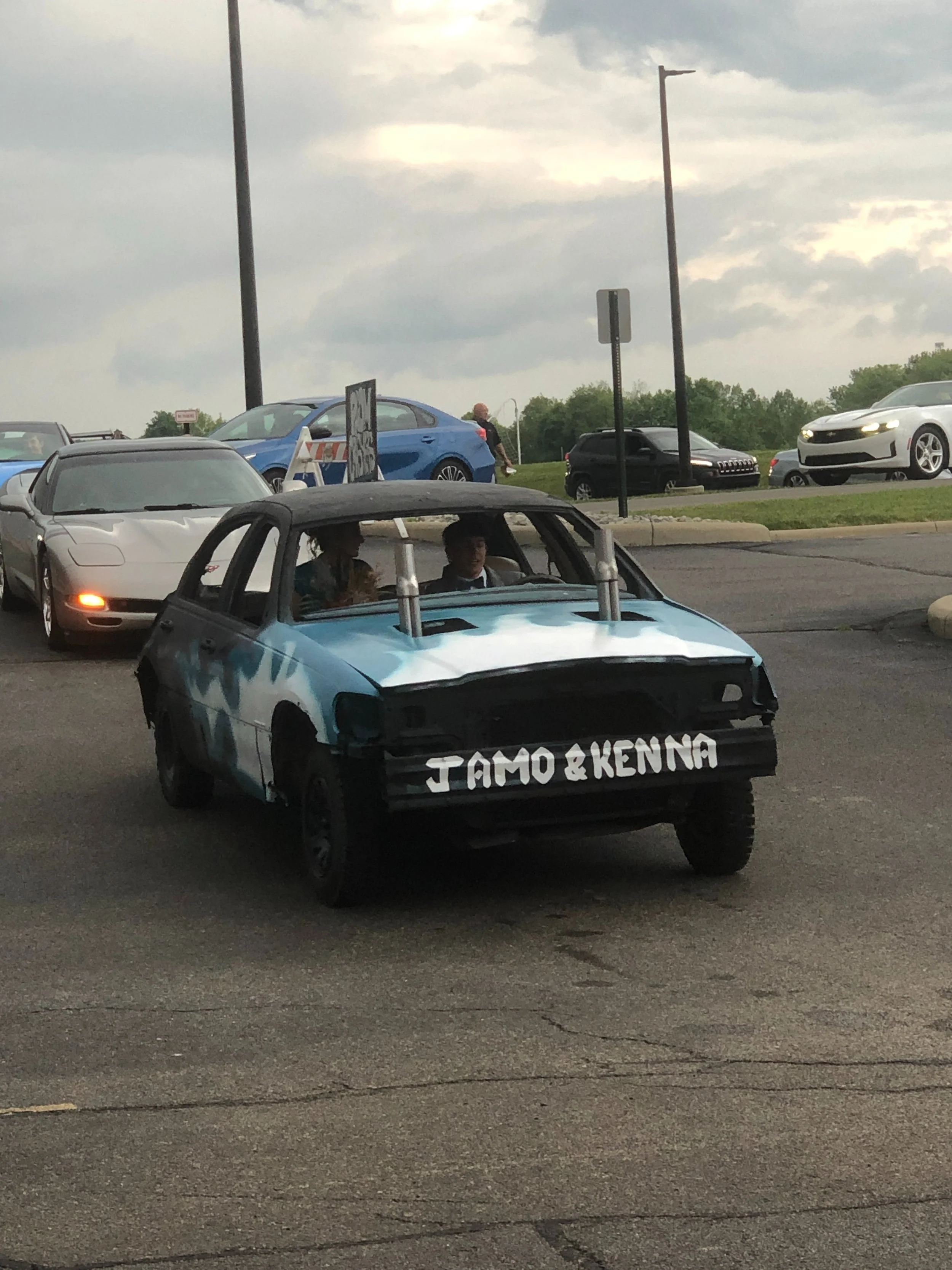 A small car with a blue and black paint job and large metal exhaust pipes, with the words 'JAMO & KENNA' painted on the front in white. Two people are sitting in the car.