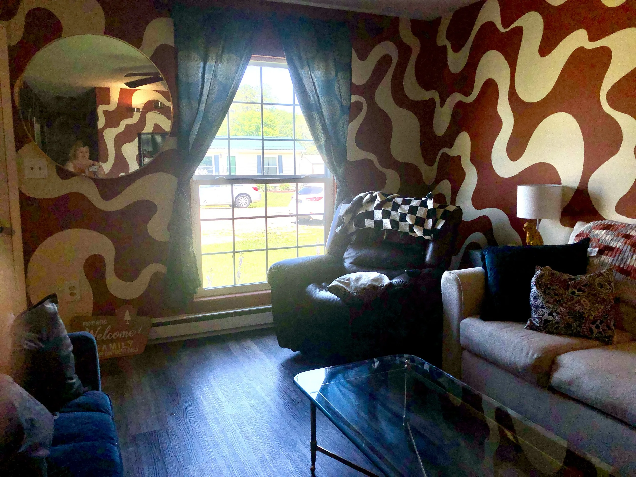 Living room with colorful wavy pattern wallpaper, large window with curtains, and furniture including a dark green armchair with a checkered blanket, a beige sofa with patterned pillows, and a glass coffee table.
