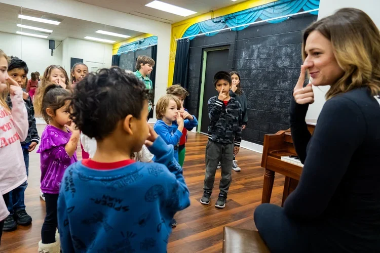 A woman appears to be talking to or teaching a group of children in a classroom or performance space, with children standing and listening to her.
