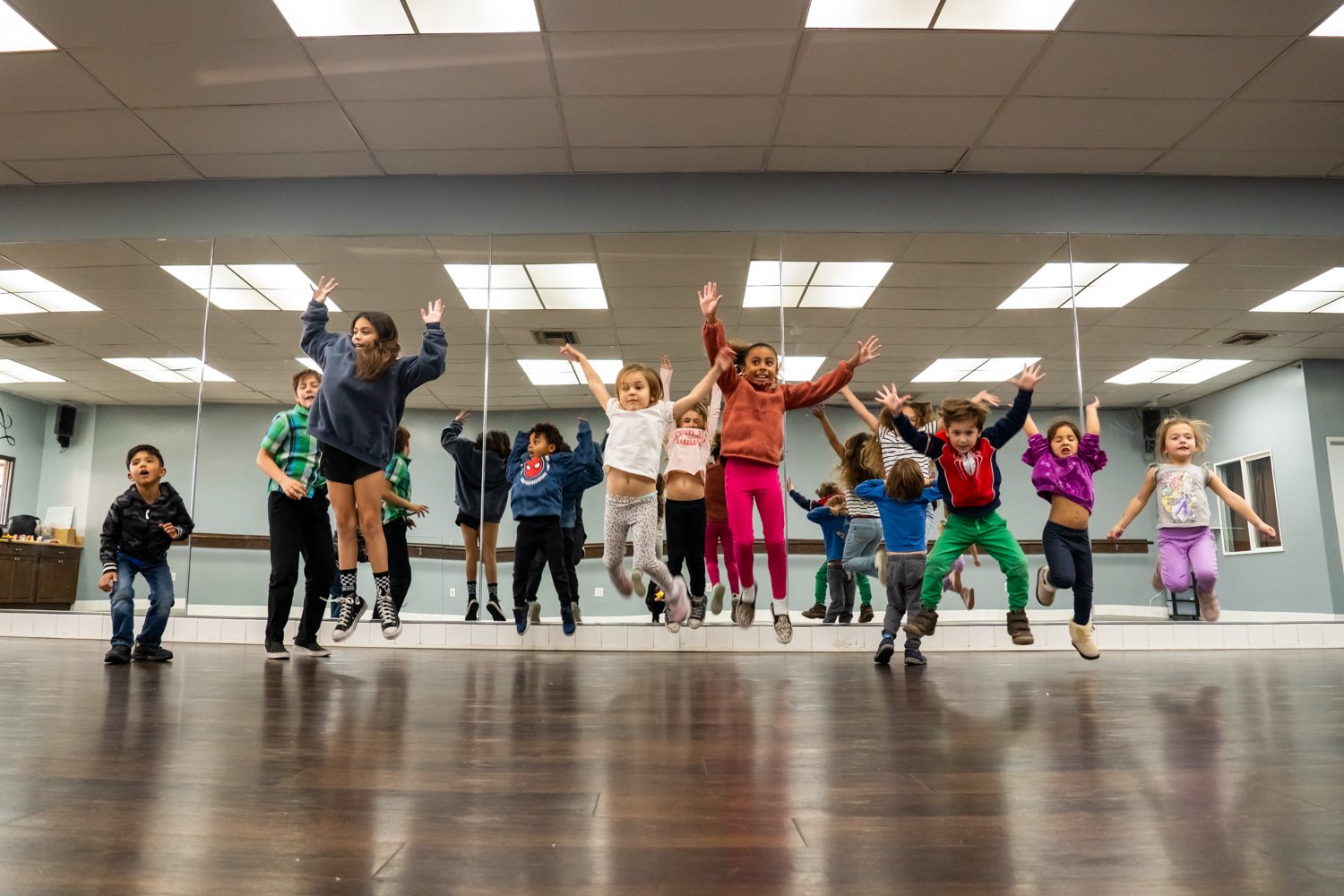 Children jumping in a dance or exercise class in a studio with a large mirror on the wall.