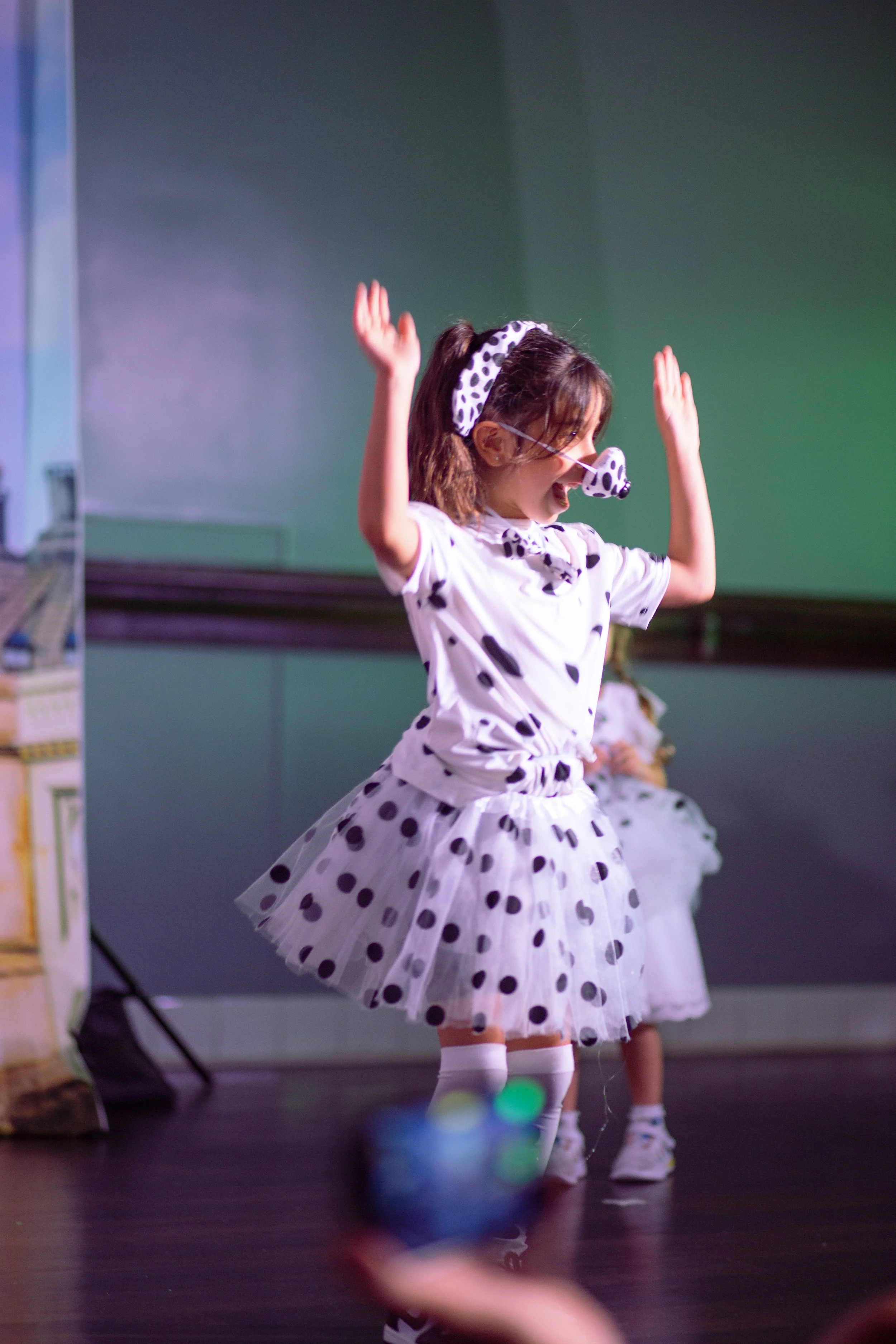 A young girl dancing on stage, dressed in a Dalmatian costume, with a matching headband and nose accessory, smiling and raising her hands in the air.