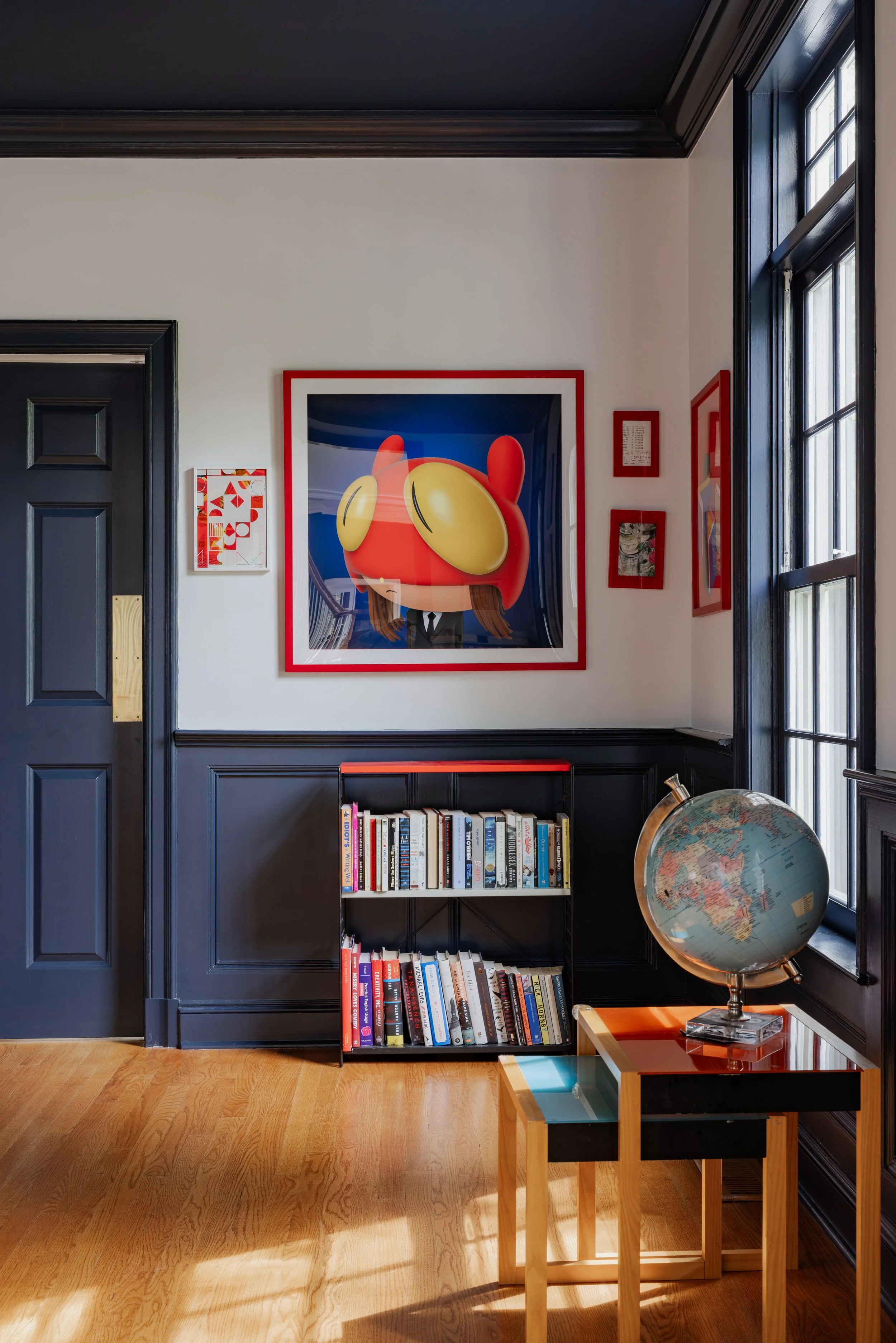 Interior view of a room with a black-made bookshelf filled with books, a globe on a small table, and artwork on the wall, with large windows letting in natural light.