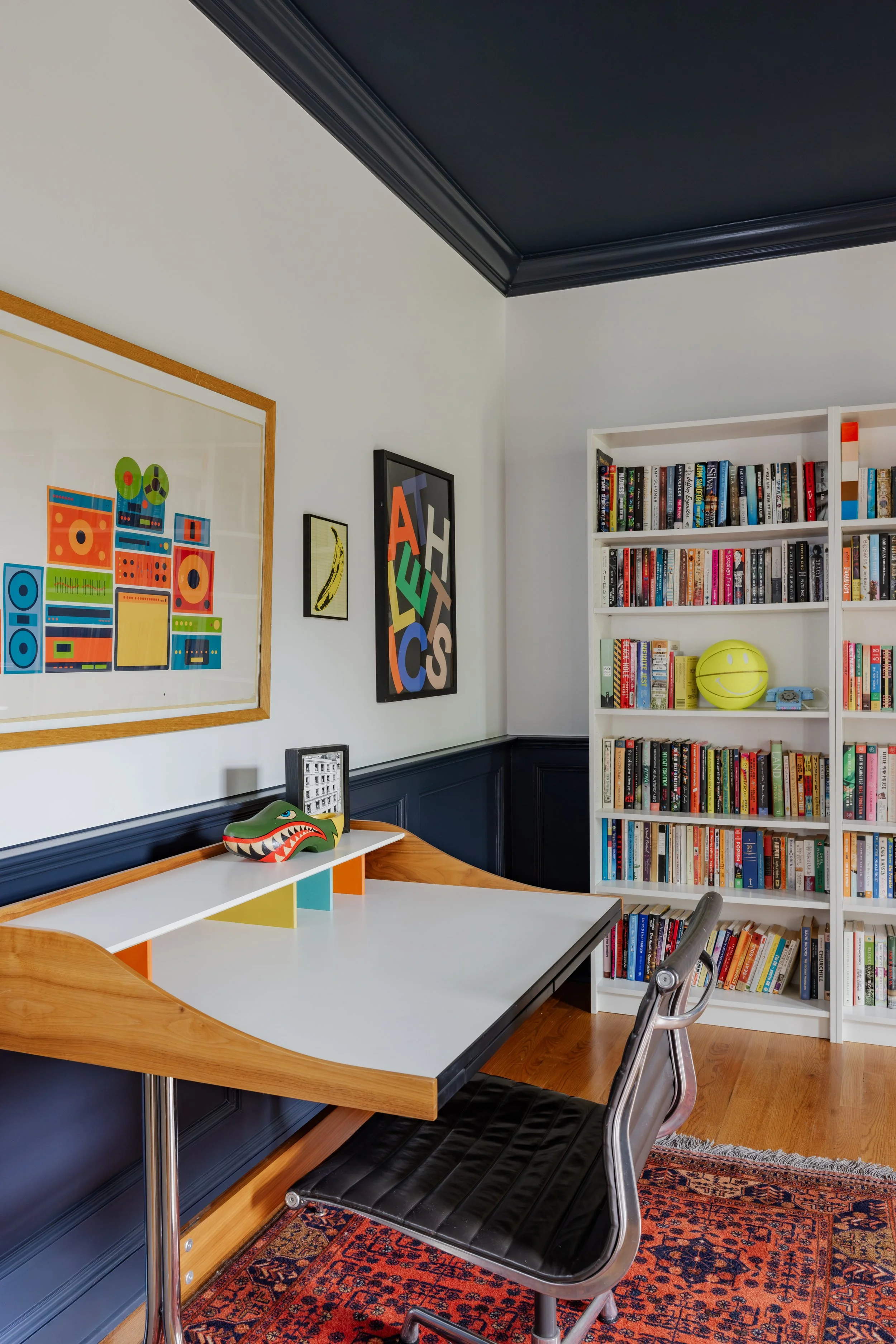 A home office with a large white desk, black chair, and a colorful carpet, decorated with framed artwork and a white bookshelf filled with books and a yellow smiley face item.