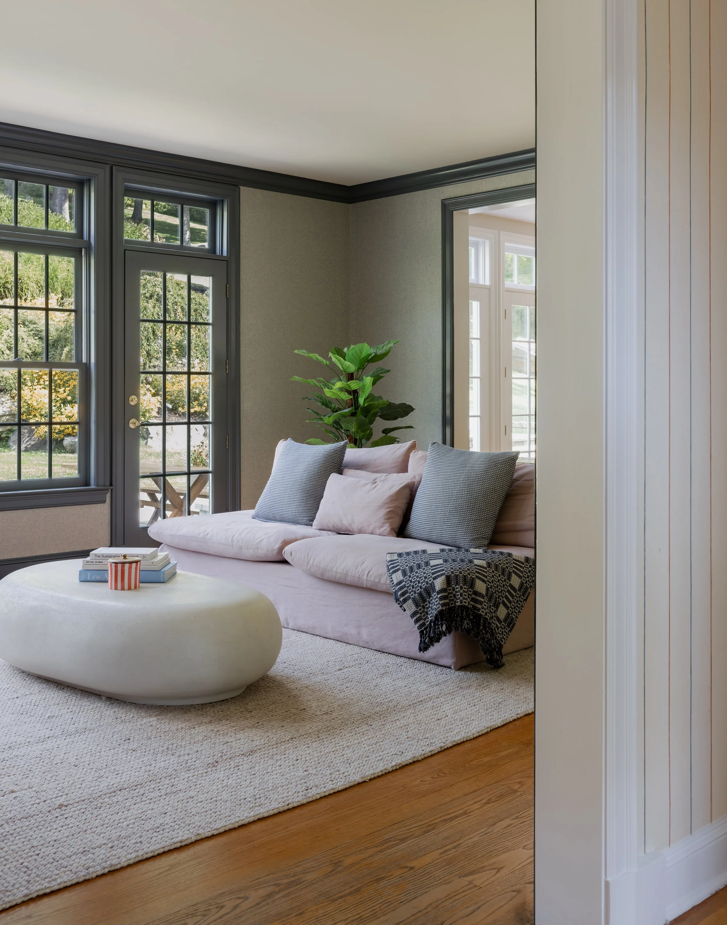 Living room with a beige sofa adorned with pillows, a round white ottoman, large windows with dark gray frames, a tall green plant, and a cream-colored wall with wood flooring.