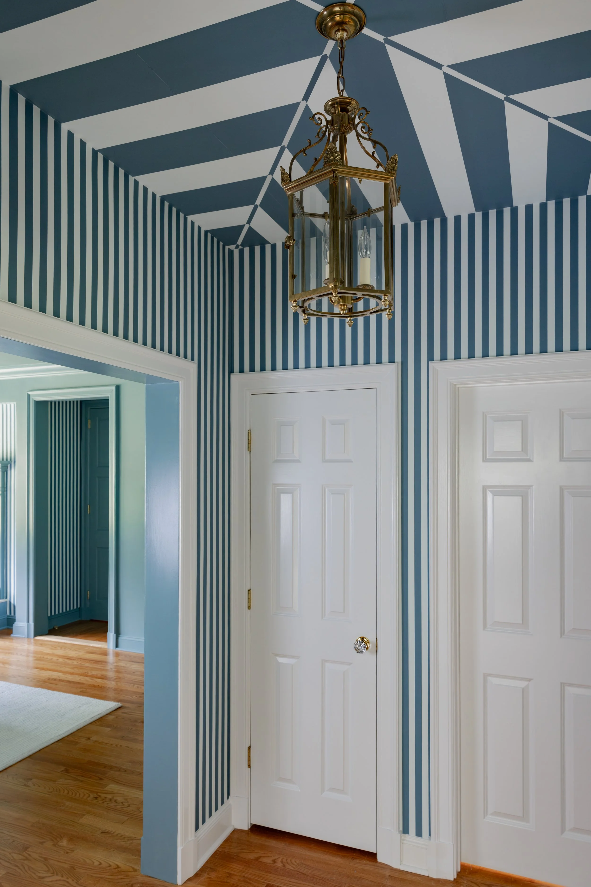 Interior view of a room with blue and white striped wallpaper on the walls and ceiling, white doors, hardwood floor, and a brass chandelier hanging from the ceiling.