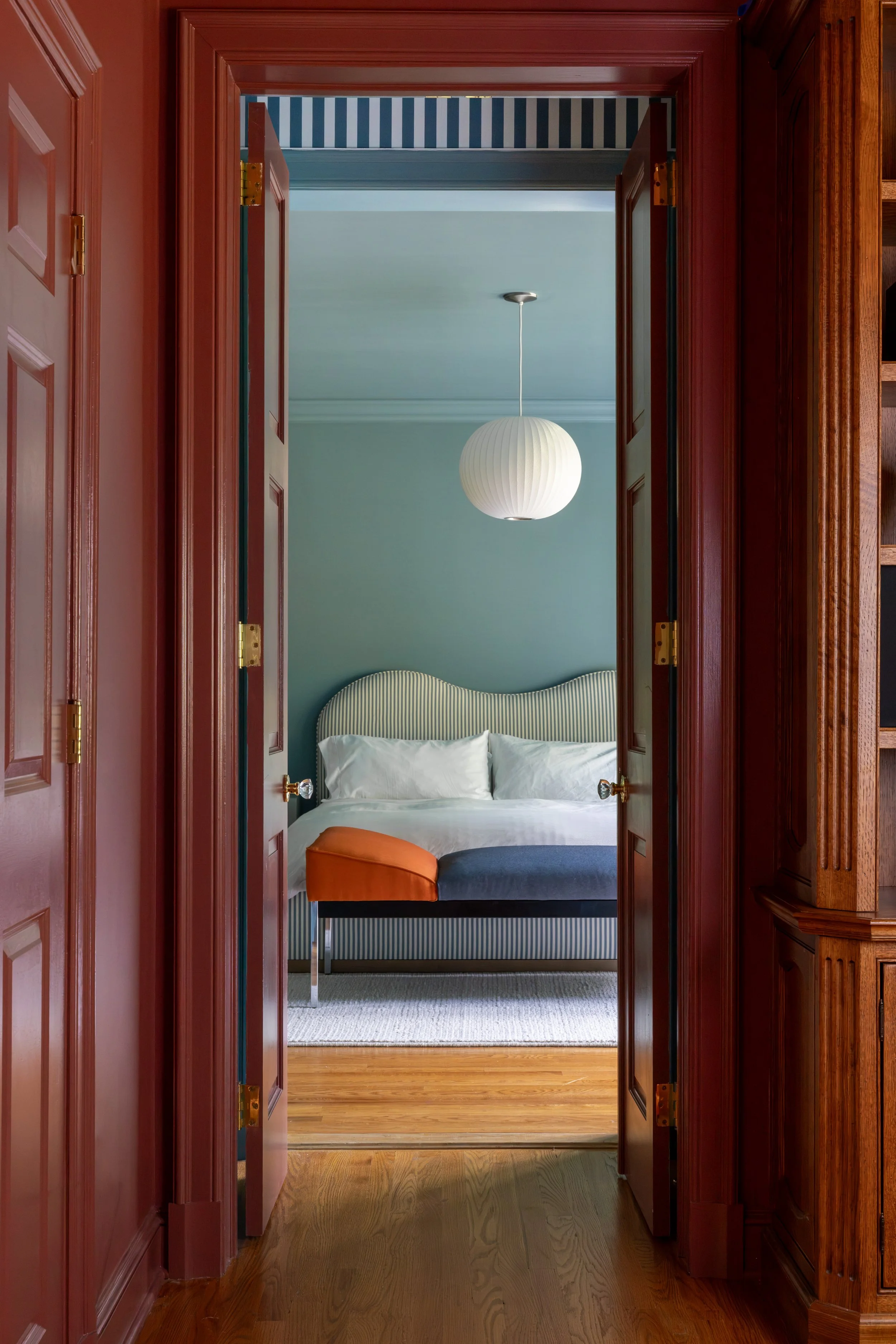 View into a bedroom through open double doors. The room has a striped headboard, a hanging round white lamp, and a small orange stool at the foot of the bed. The flooring is wooden.