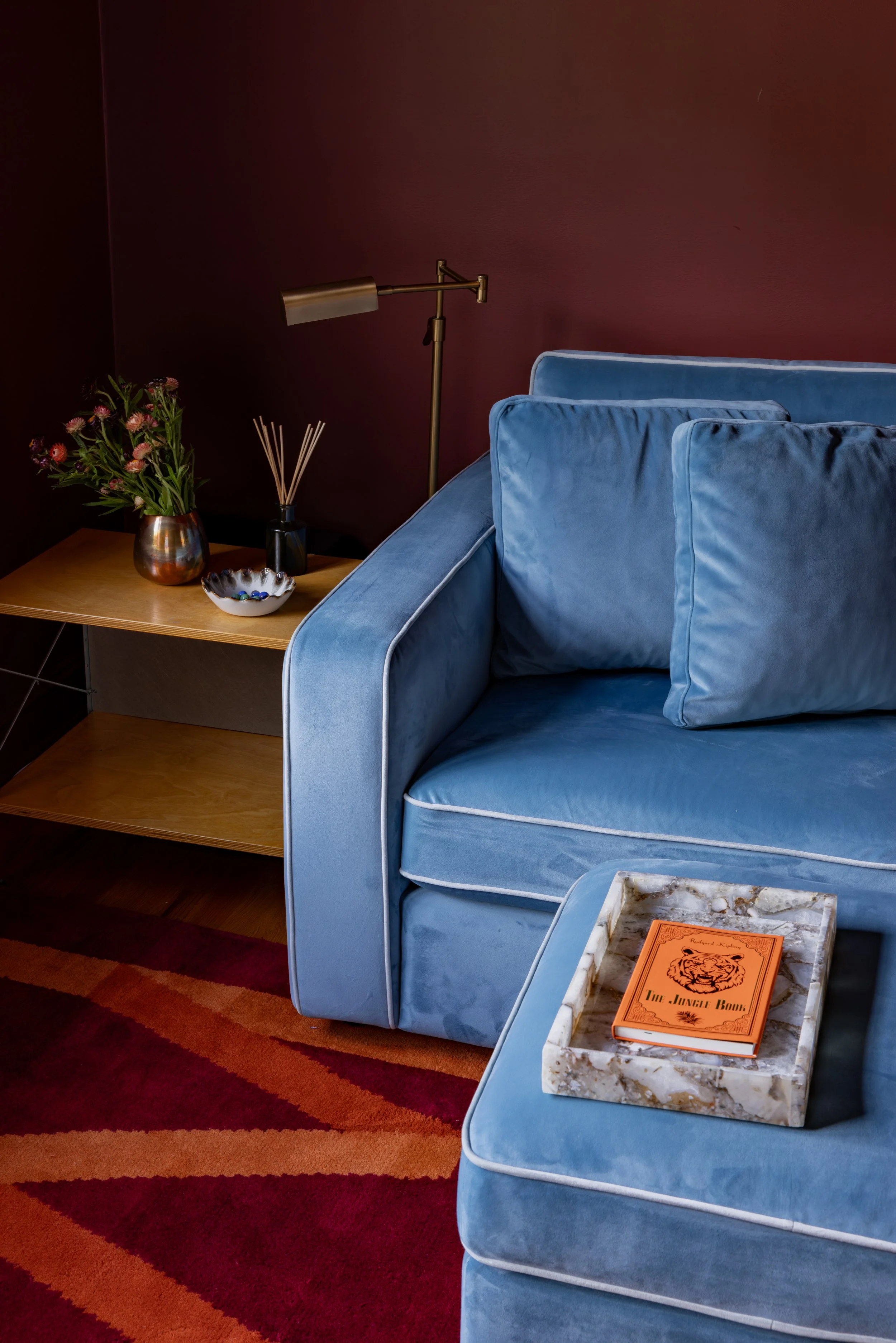 A blue velvet sofa with pillows, a marble tray with an orange book titled "The Jungle Book," a wooden side table with a flower vase, a bowl, and a reed diffuser, and a brass reading lamp against a dark red wall.