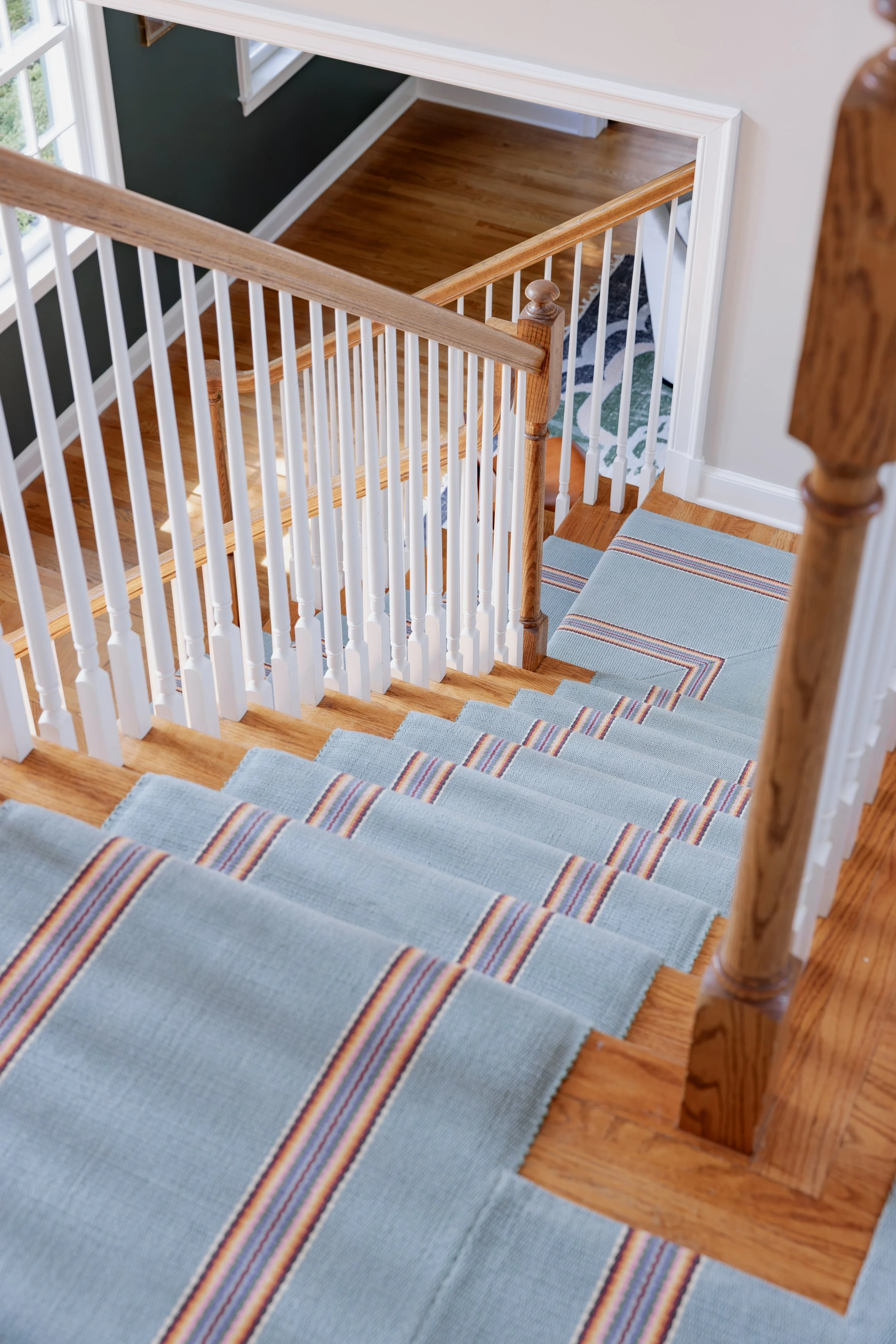 A staircase with a blue runner carpet and a wooden banister. The stairs go down to a landing, and there are windows on the left side of the image.