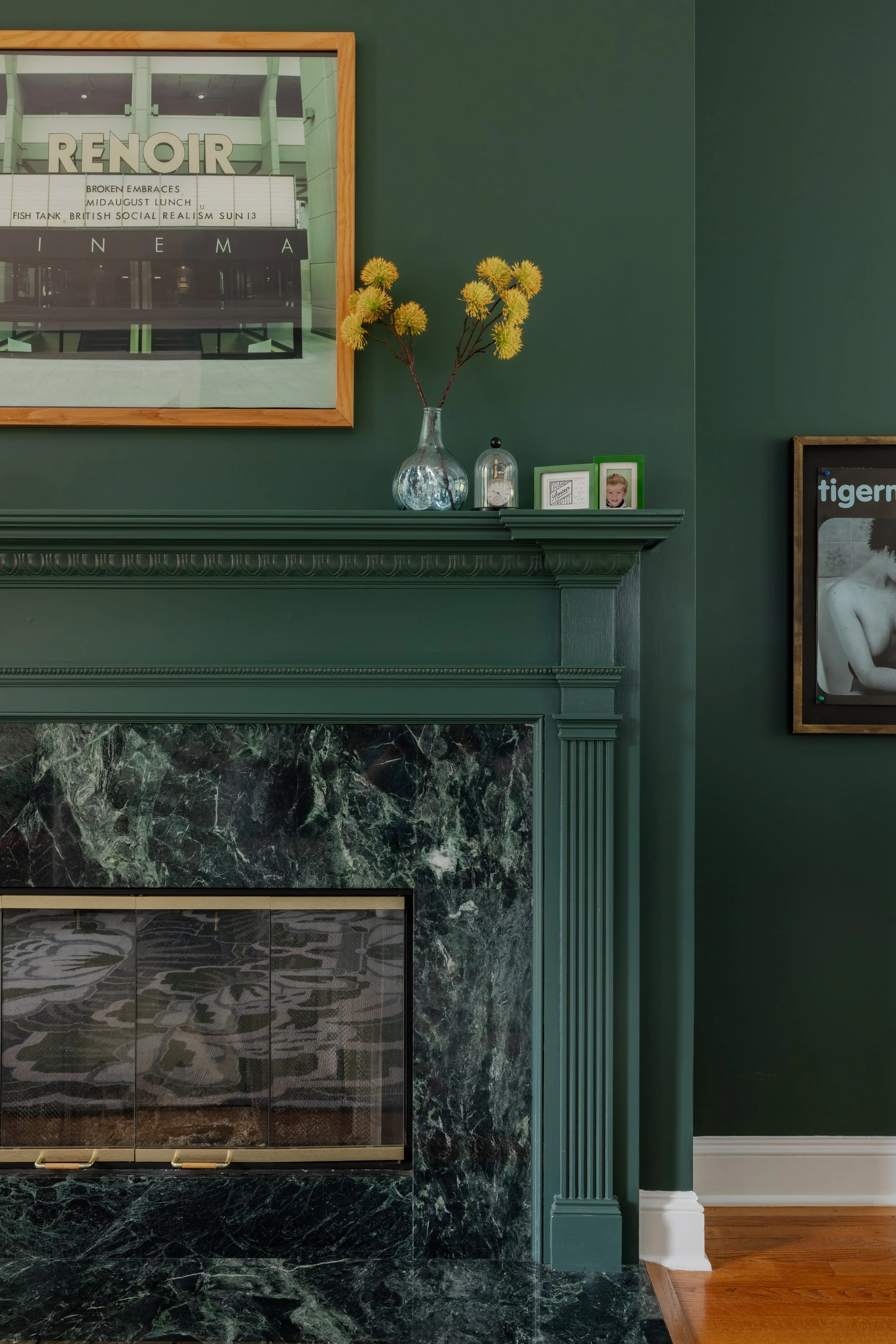 Close-up of a fireplace mantel painted dark green, topped with a clear glass vase holding yellow-green flowers, framed photographs, and a small decorative item, against a green wall with a framed art piece on the right.