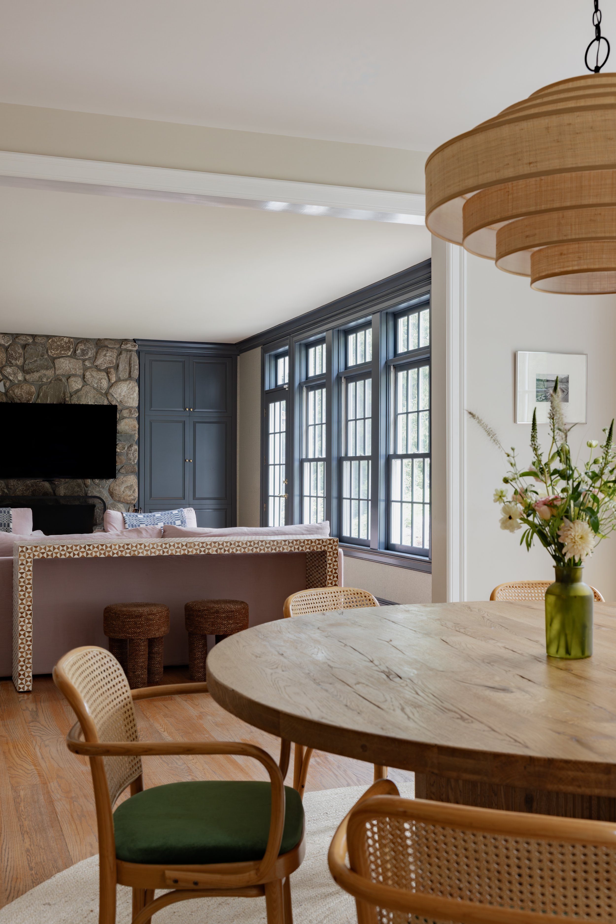 Interior view of a living and dining room with large windows, a stone fireplace, built-in blue cabinets, a round wooden table with chairs, floral arrangement in a green vase, and a wooden ceiling lamp.