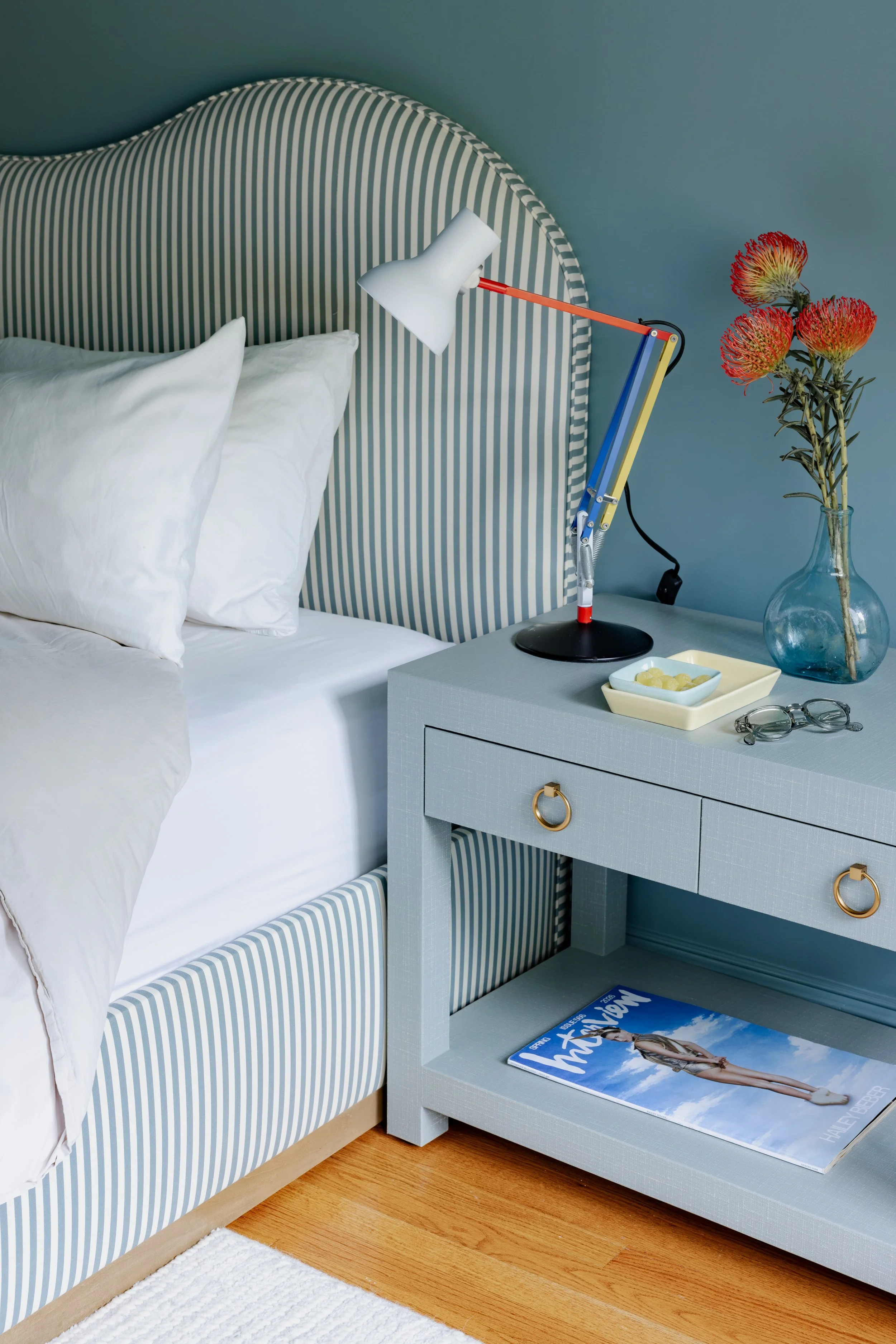 A bedroom with a blue and white striped headboard, a white bedside table, and a vase with orange flowers. There is a white bed with white pillows and a magazine on the floor.