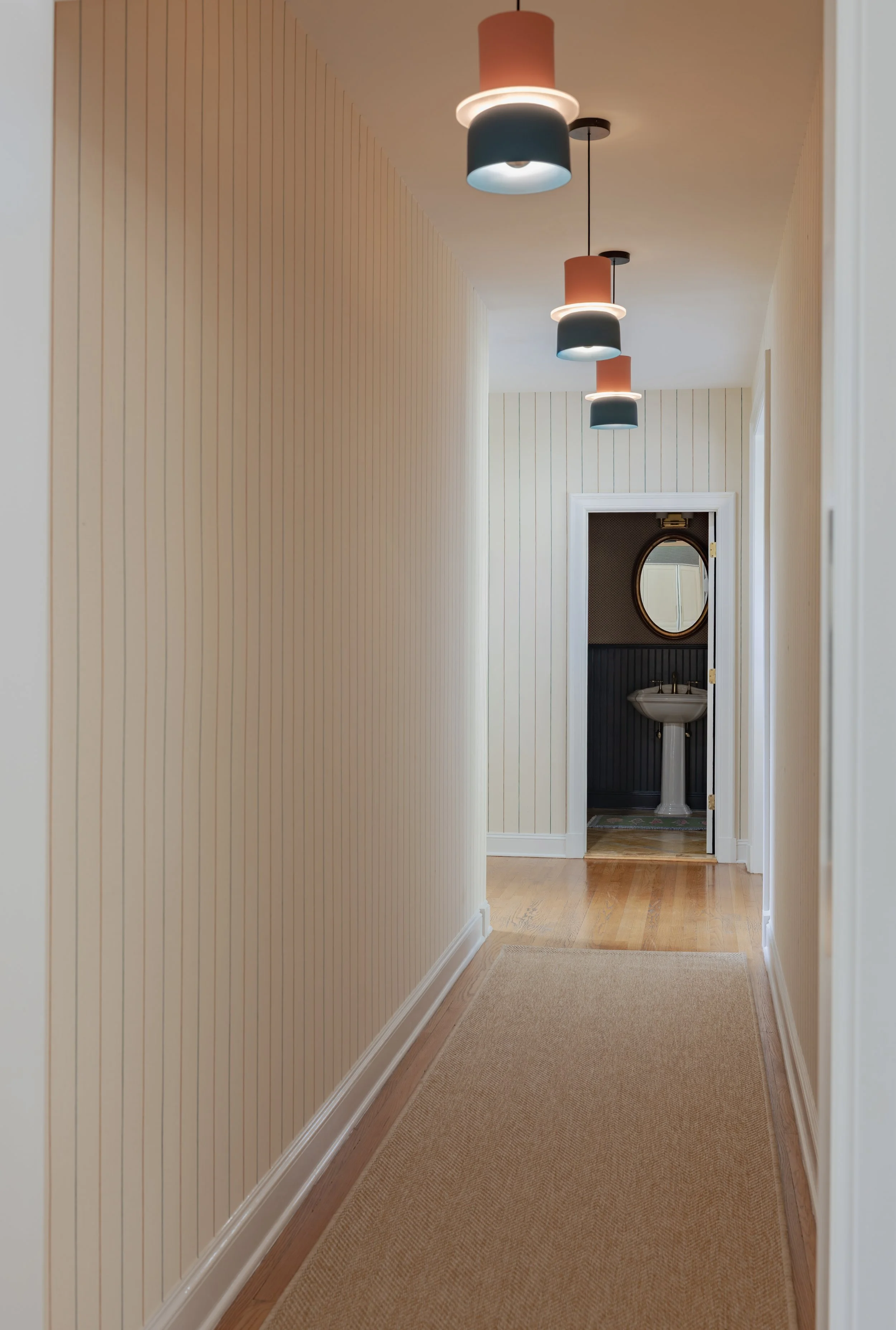 A hallway with beige striped wallpaper, a beige carpet runner, and three hanging pendant lights with peach and dark gray shades. At the end of the hallway, there is a doorway leading to a small room with dark wainscoting, a white pedestal sink, and a