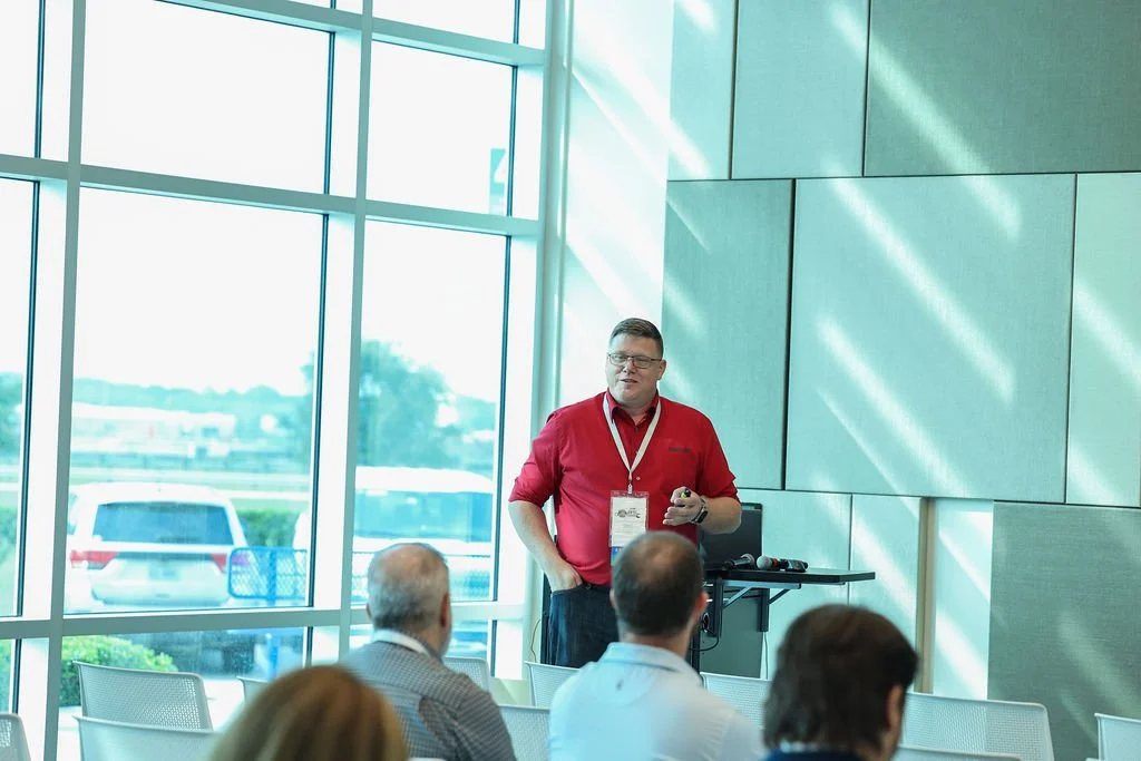 Man giving a presentation to a seated audience in a modern room with large windows and sunlight.