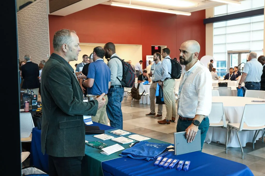 Two men are engaged in conversation at a conference or trade show, standing over a table with promotional items like pens and brochures. One man has gray hair and a beard, wearing a dark blazer, while the other is bald with a beard, wearing a white s